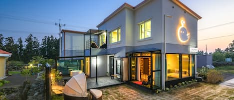 A modern two-story house with a glass facade, a balcony, and a well-lit interior.