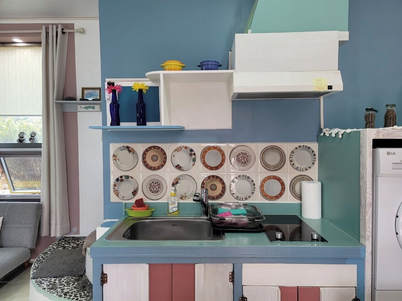 A compact kitchen with a sink, a stove, and a microwave. There are decorative plates on the wall and a small shelf with kitchen items.