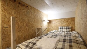 A small, cozy room with a wooden wall, a bed with a plaid bedspread, and a wall-mounted light fixture. - Odori Backpackers (Sapporo)