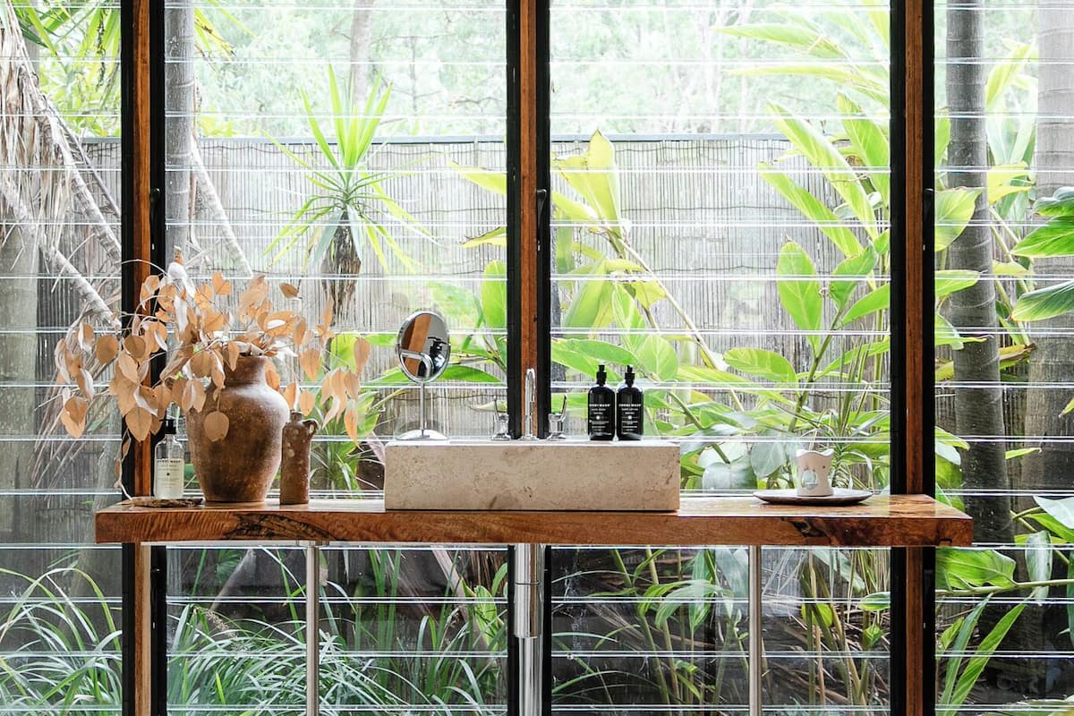 Une chambre avec une grande fenêtre, une table de chevet ornée d’un vase et un lit recouvert d’une couverture blanche.