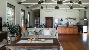 A spacious living room with a large sofa, wooden coffee table, and potted plants. A kitchen with modern appliances is visible in the background. - The Boathouses at Leaves & Fishes (Lovedale)