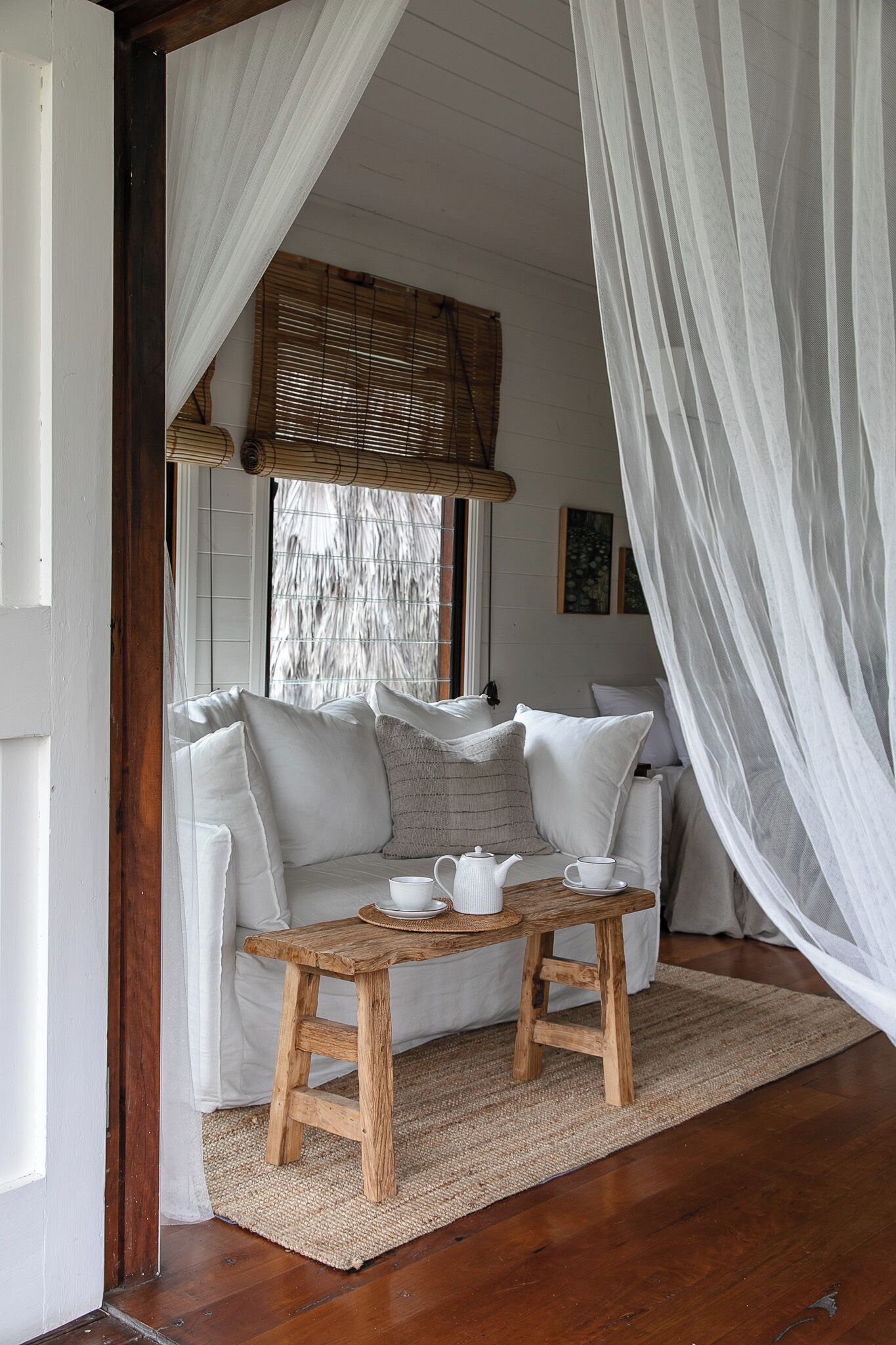 A cozy room with a wooden bench, white cushions, a woven ceiling, and a large window with blinds.