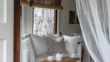 A cozy room with a wooden bench, white cushions, a woven ceiling, and a large window with blinds.