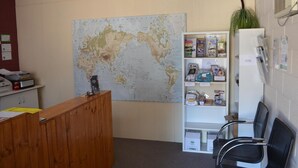 Uma área de recepção com um grande mapa-múndi, balcão de madeira, estante branca com folhetos informativos e cadeiras pretas.