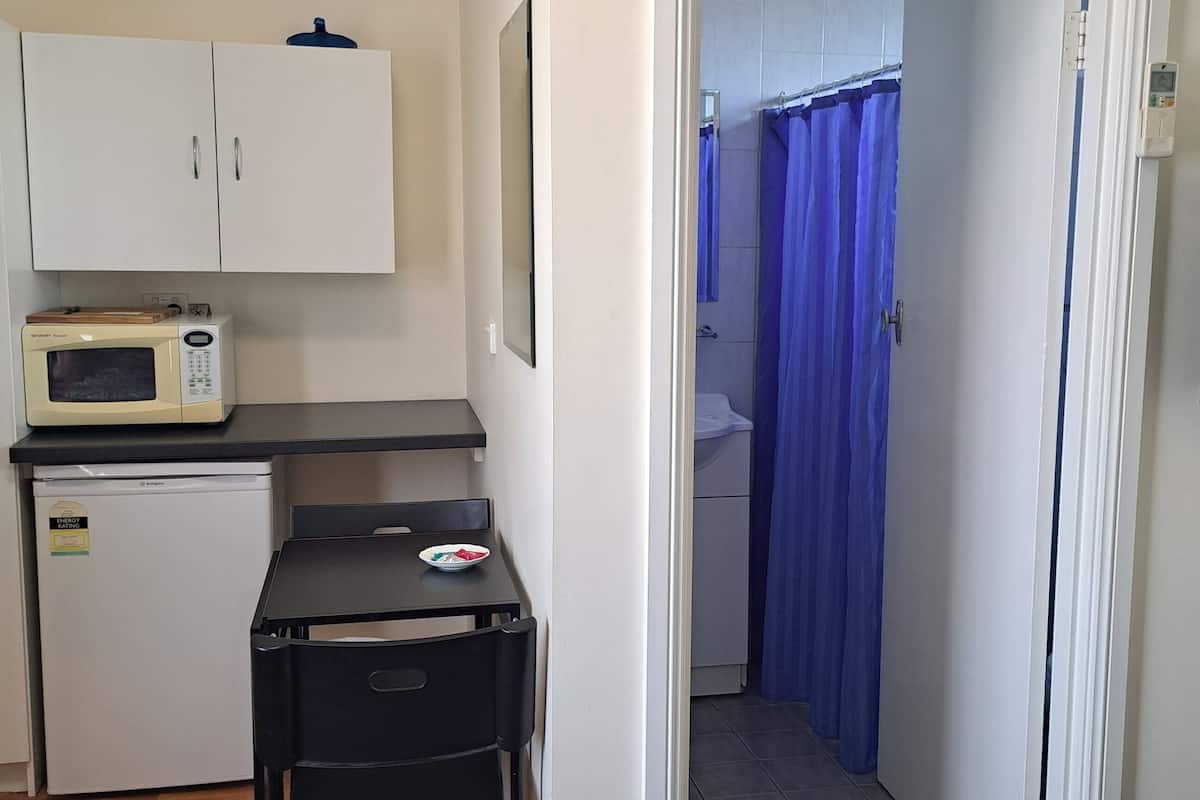 A compact kitchen with a microwave, refrigerator, and a small table with a bowl. A shower area is visible through a curtain, and a TV is mounted on the wall.