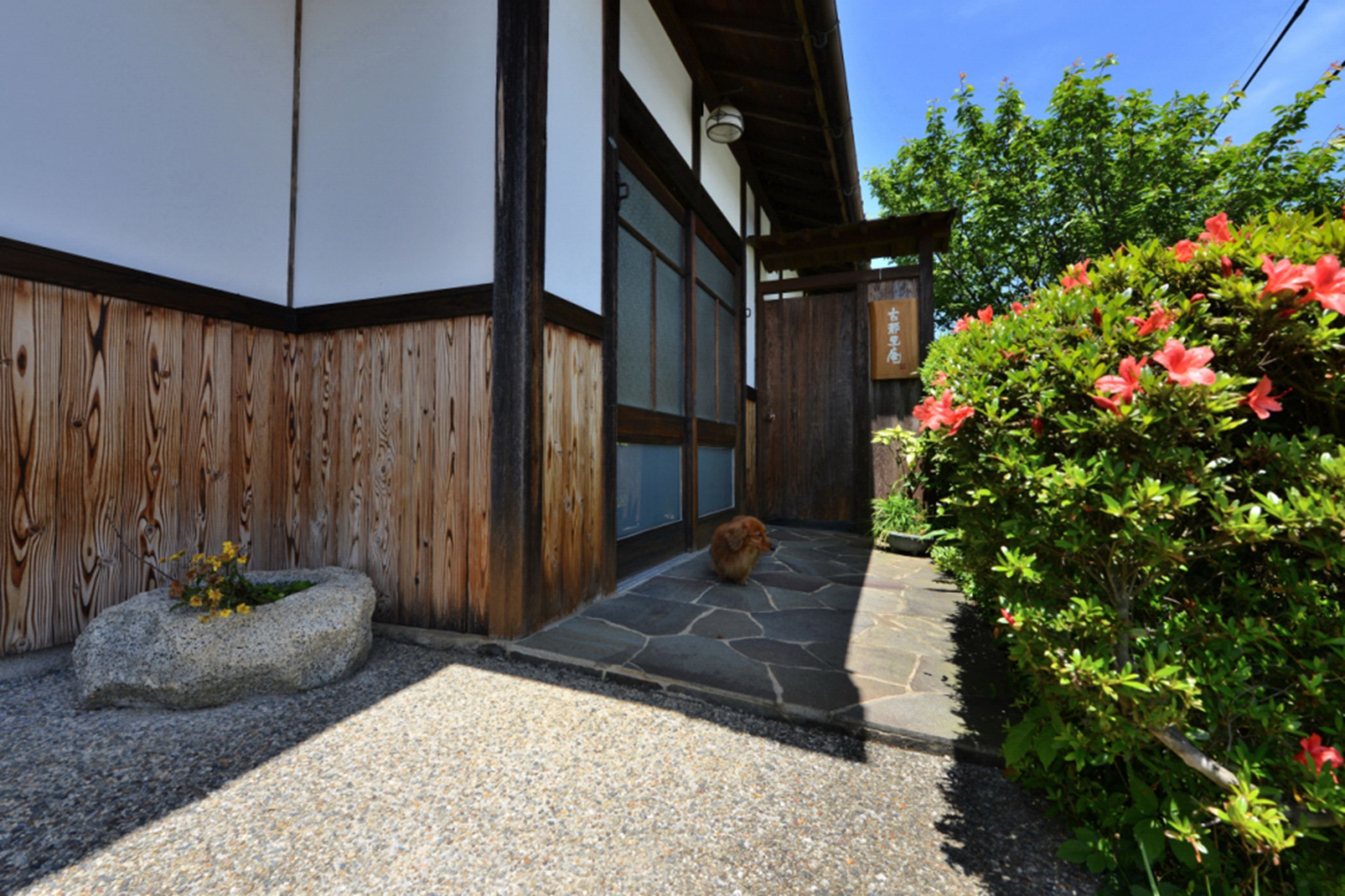 En traditionell japansk byggnad med en träfasad, en entré av sten och en trädgård med röda blommor.