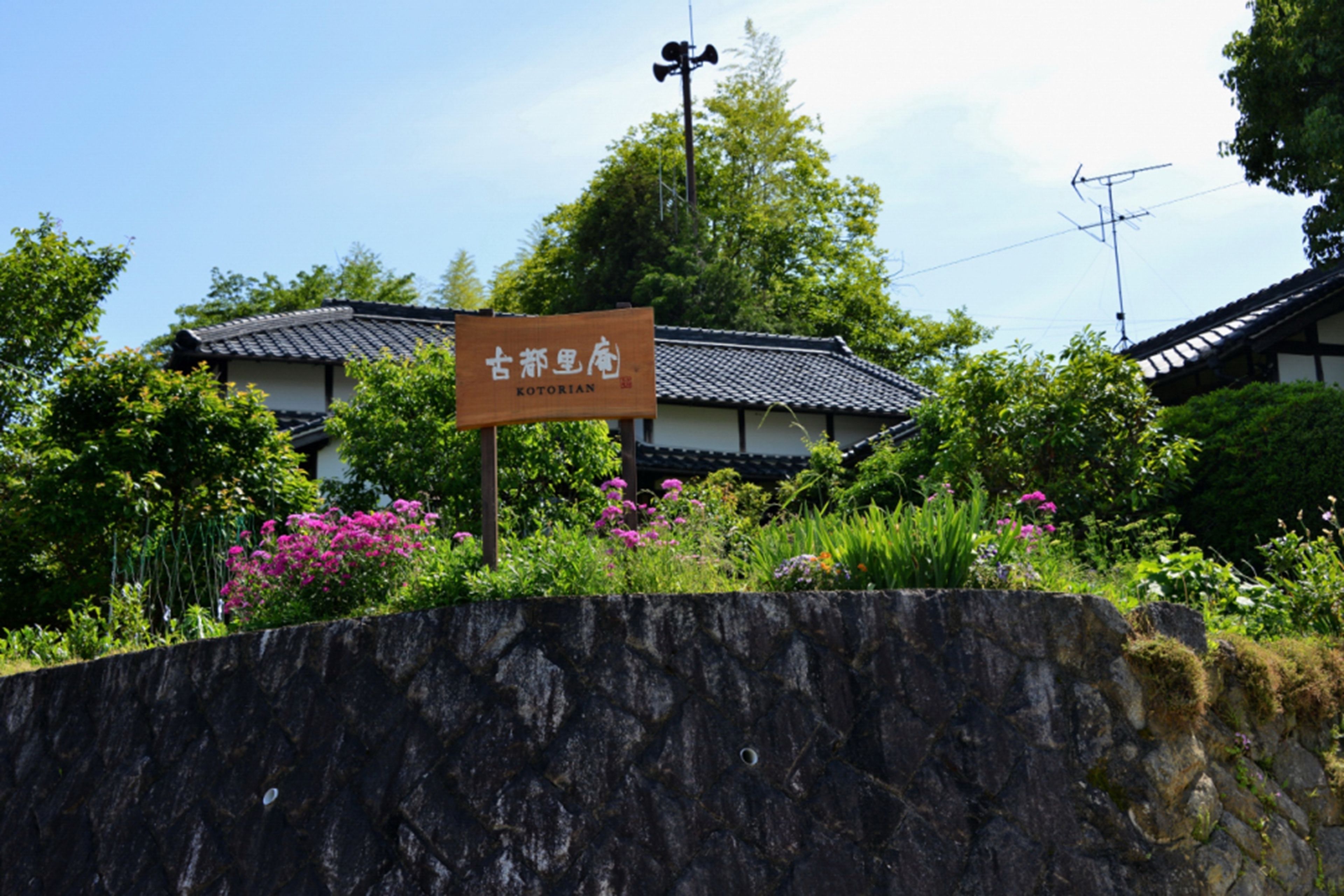 Un edificio japonés tradicional con un letrero, rodeado de abundante follaje y plantas en flor.