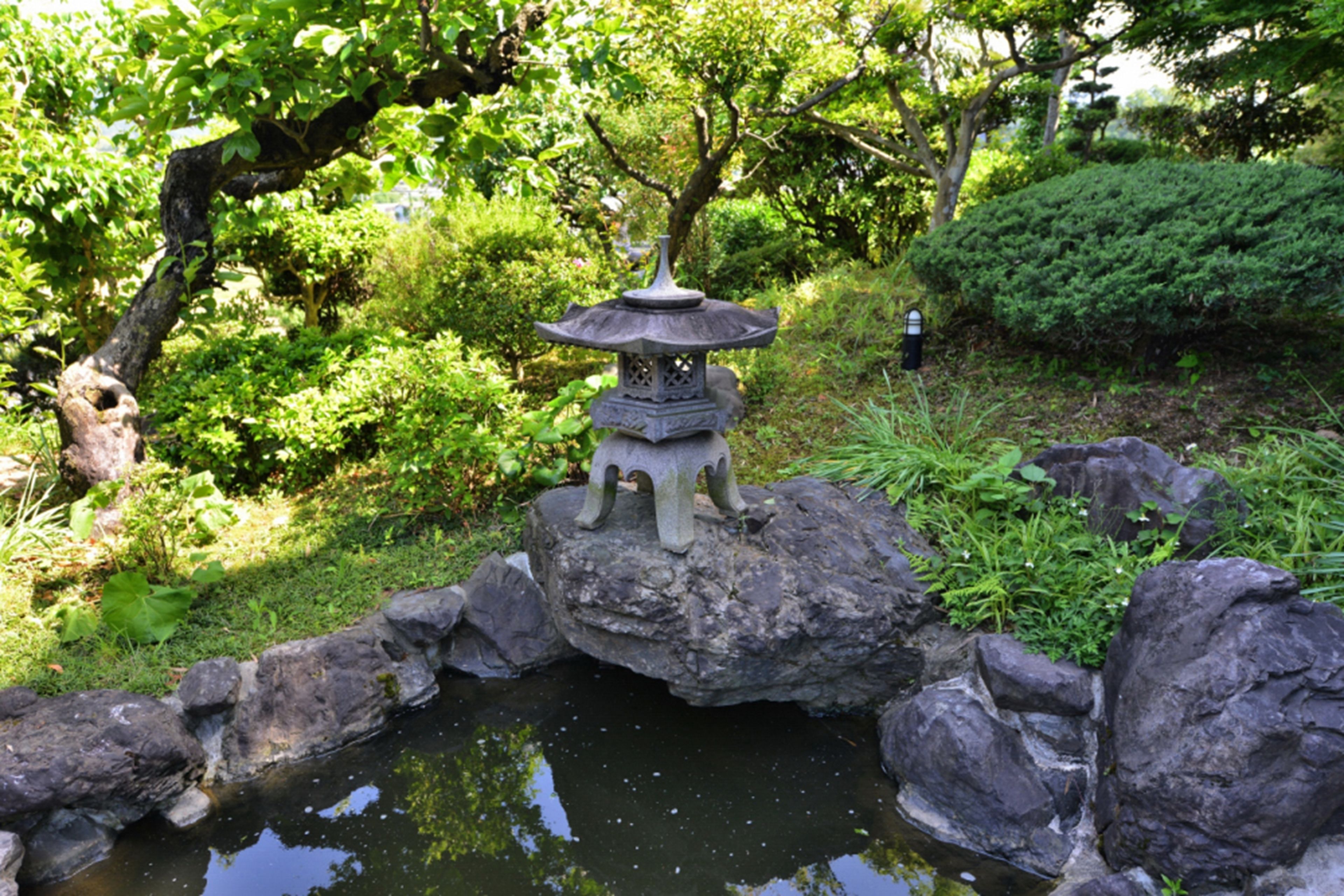 A traditional Japanese stone lantern on a rock by a pond, surrounded by lush greenery and trees.