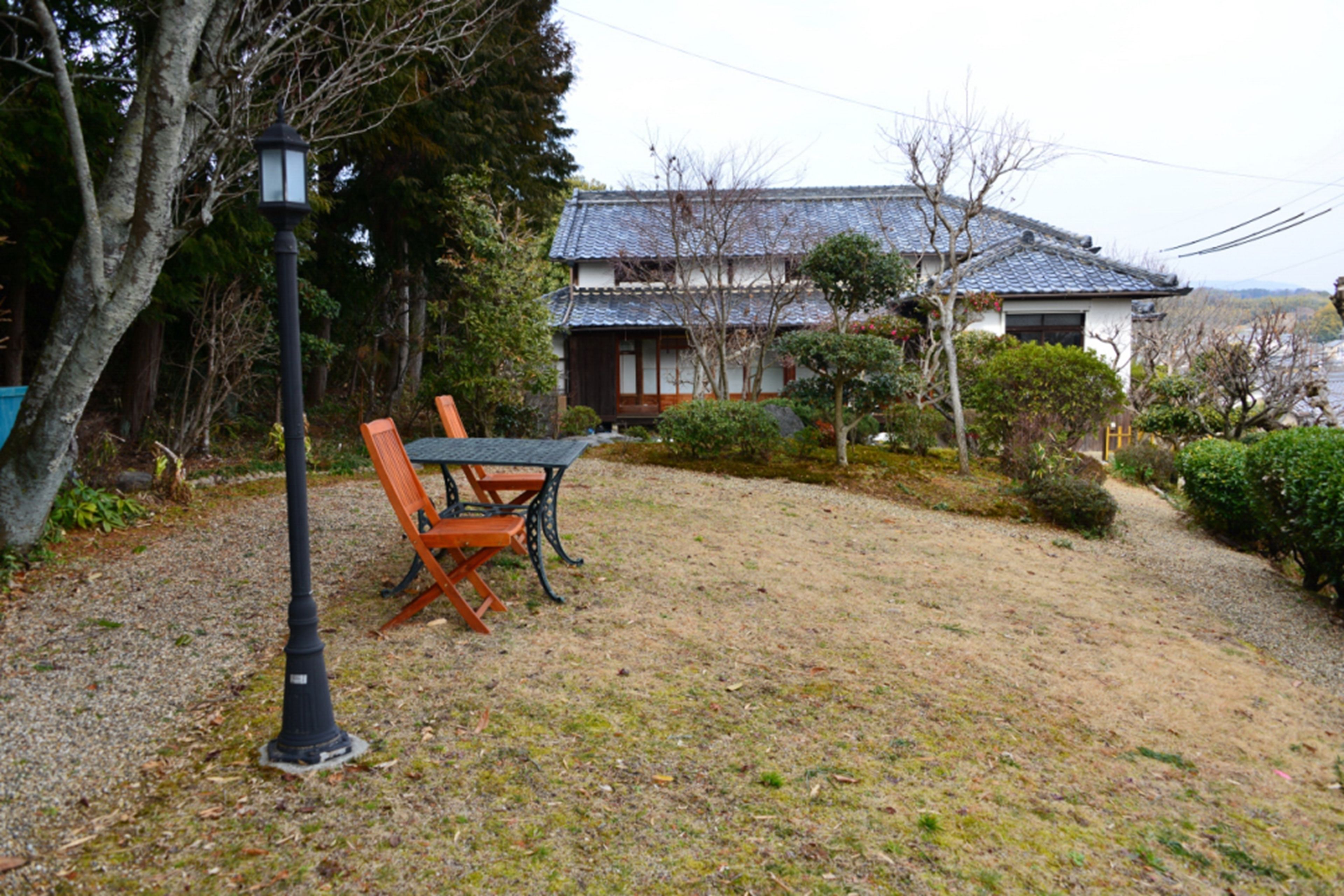 A traditional Japanese house with a tiled roof, surrounded by a gravel path and a wooden table with chairs.