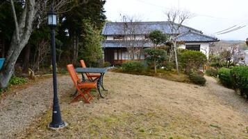 A traditional Japanese house with a tiled roof, surrounded by a gravel path and a wooden table with chairs.