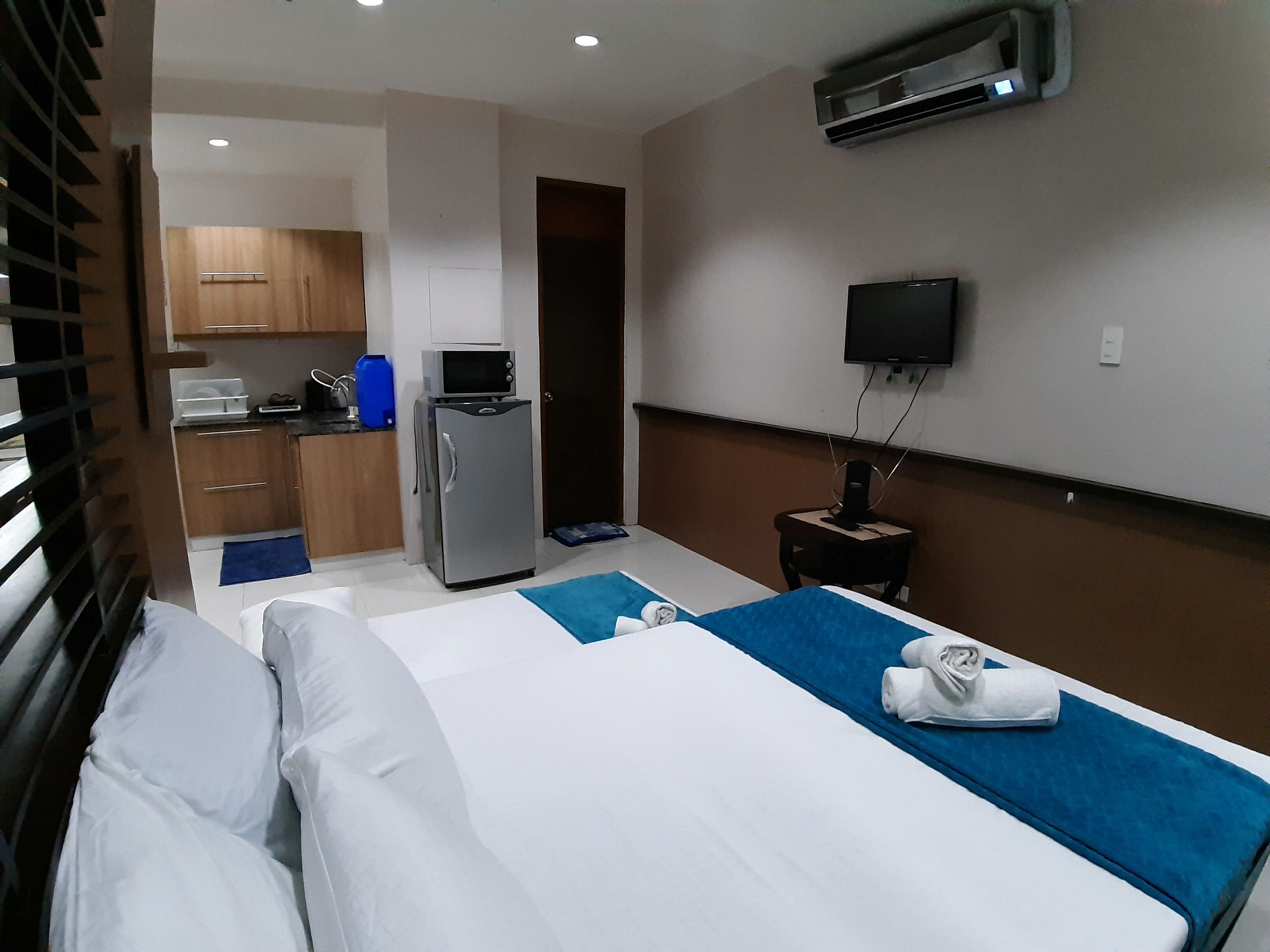 A hotel room with a bed, a small kitchenette, a microwave, a refrigerator, a TV, and an air conditioning unit.