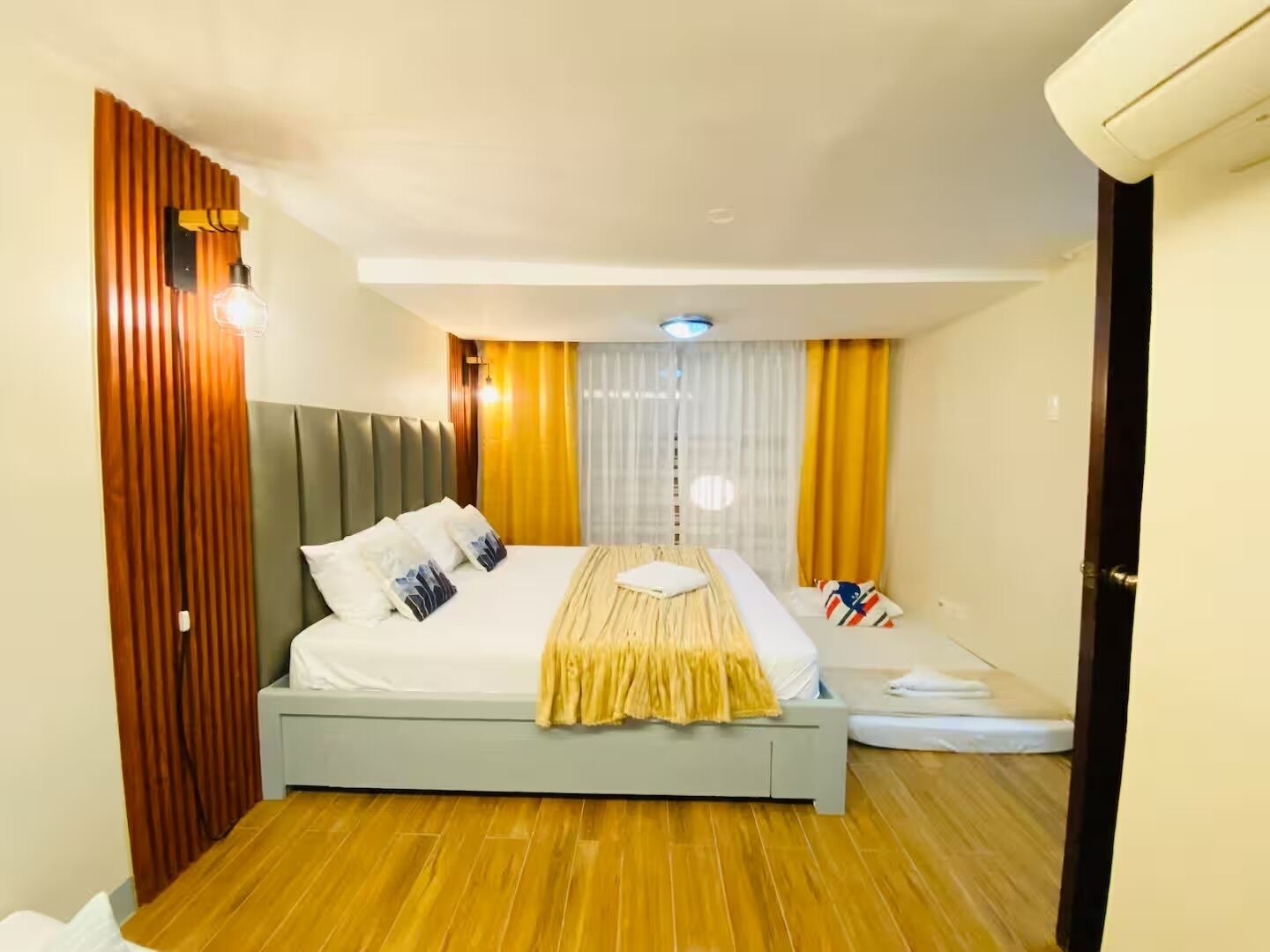 Une chambre d’hôtel avec un lit, une table de chevet, une fenêtre avec des rideaux et un panneau en bois sur un des murs.