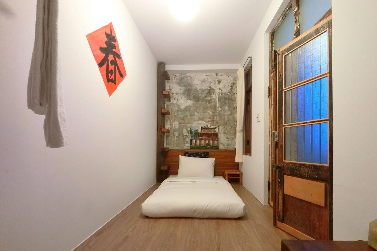 A room with a bed, a wooden floor, a wall with a painting, and a door with blue-tinted glass panels.