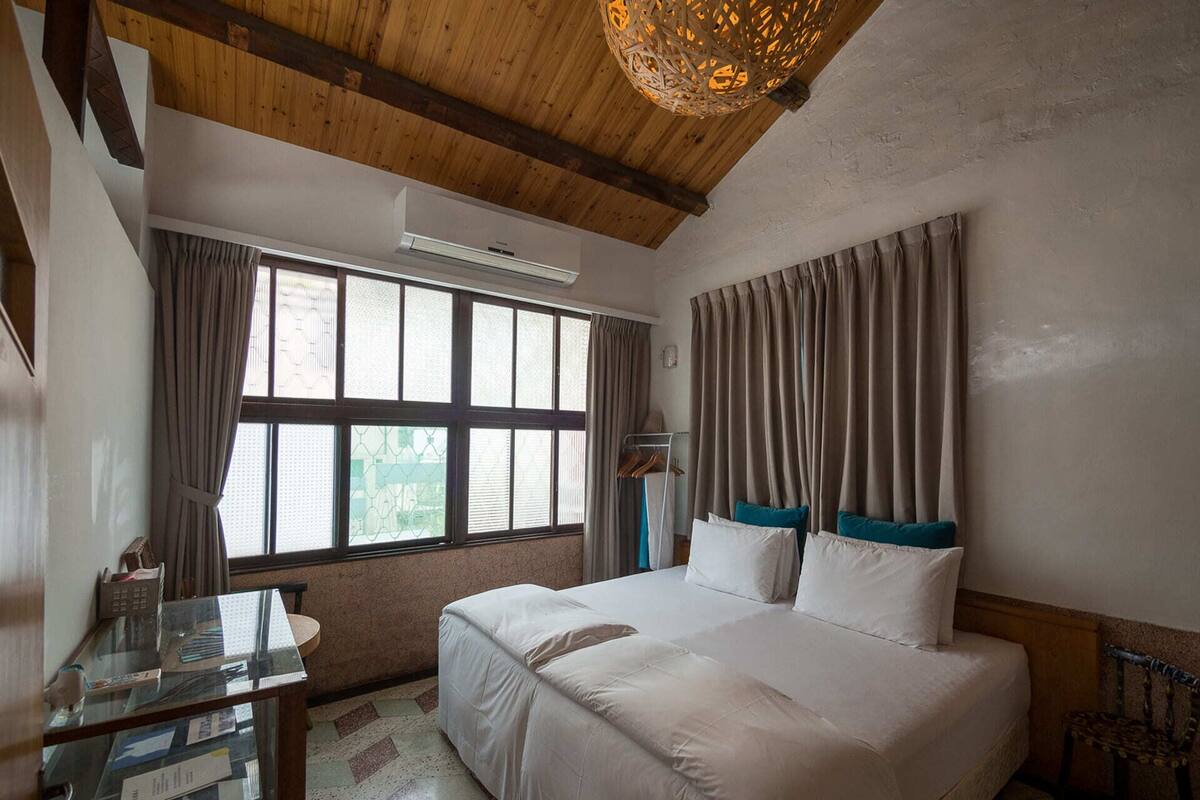A bedroom with a large bed, a desk with papers, a window with curtains, and a wooden ceiling.