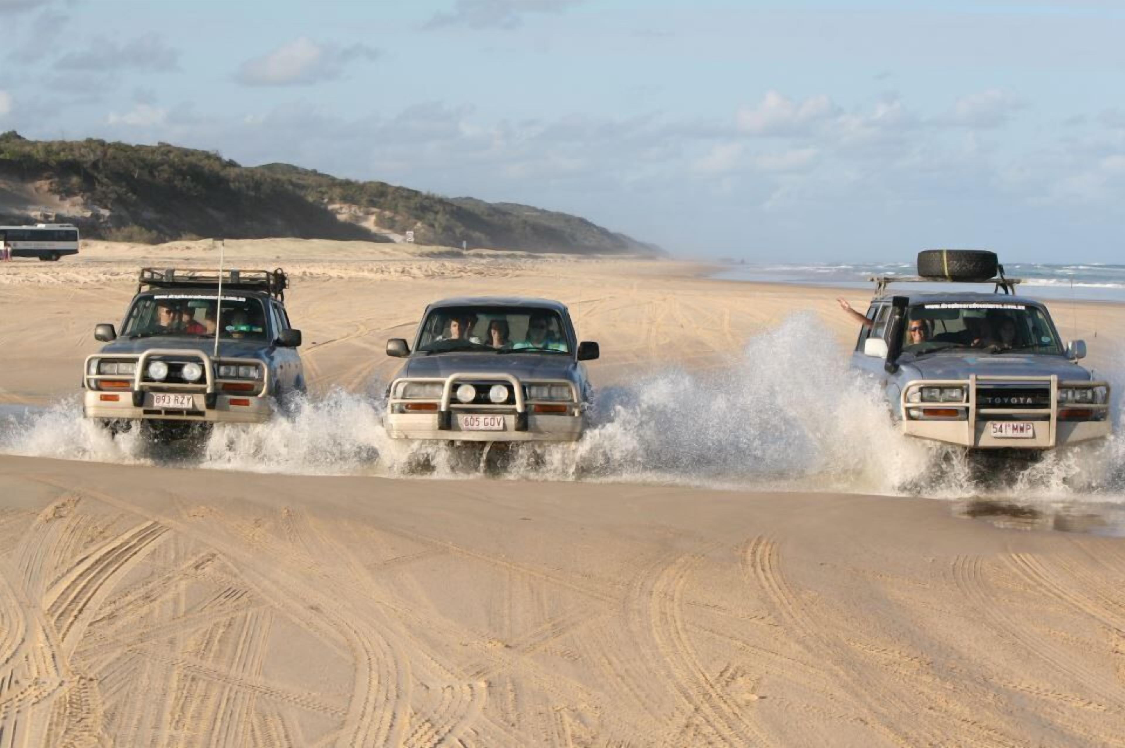 Three off-road vehicles driving through a sandy area with a beach and ocean in the background.