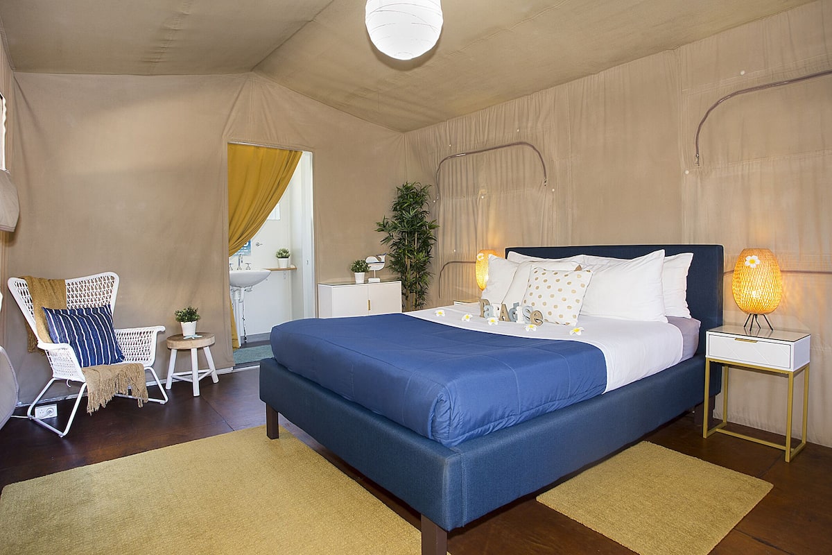 A bedroom with a large bed, bedside tables, a chair, a small table with a plant, and a window with curtains.