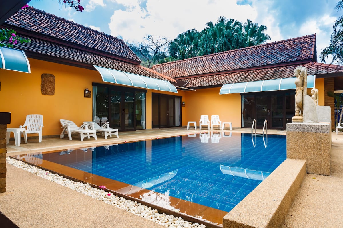 A swimming pool with lounge chairs and a statue, surrounded by a building with a tiled roof and yellow walls.