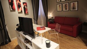 A modern living room with a red sofa, a white table with a remote control, a book, and a candle. There are framed pictures on the wall and a window with curtains. - K Rooms (Rome)