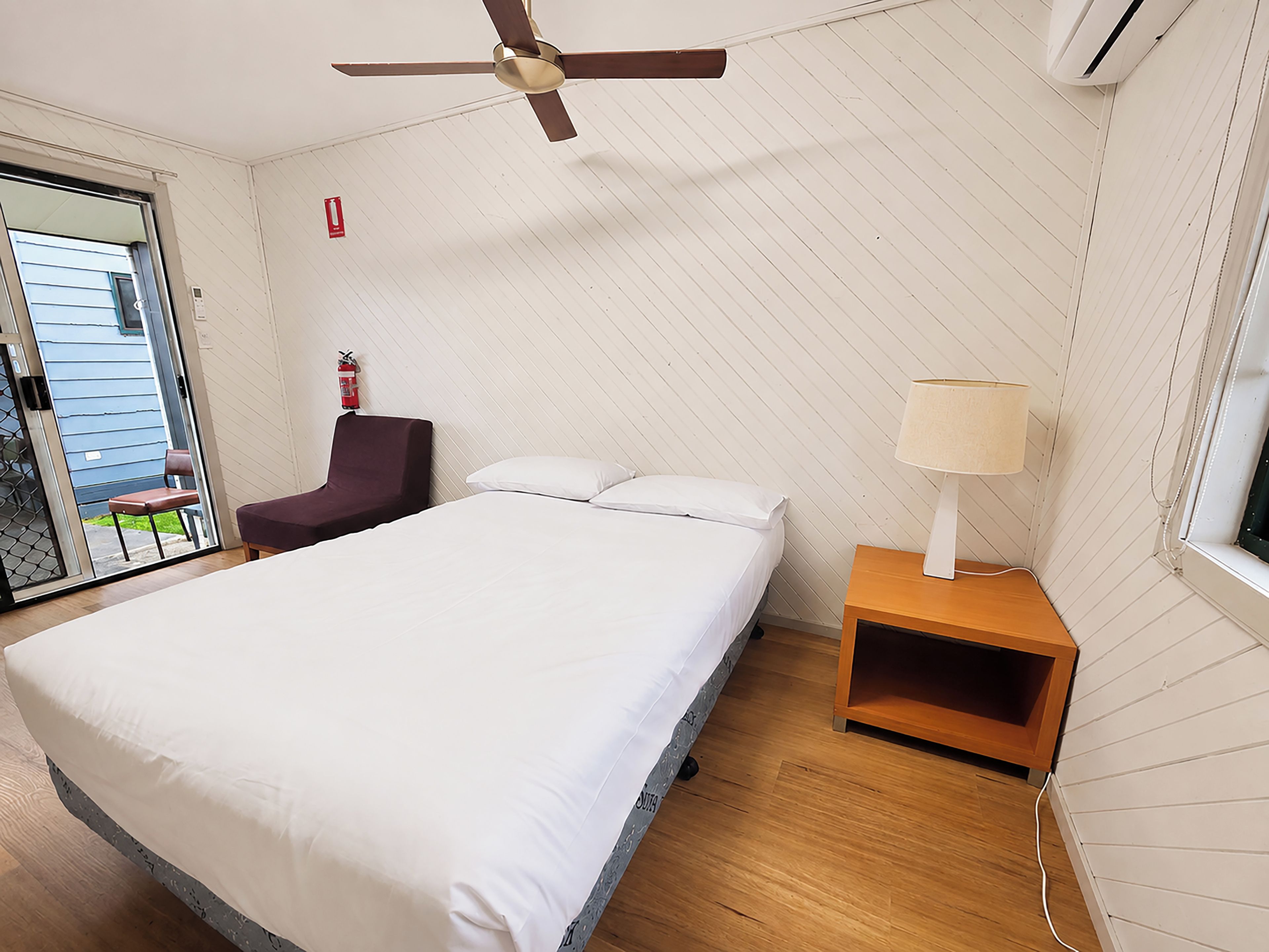A hotel room with a bed, a chair, a bedside table with a lamp, a ceiling fan, and a fire extinguisher.