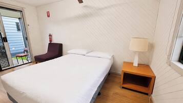 A hotel room with a bed, a chair, a bedside table with a lamp, a ceiling fan, and a fire extinguisher.