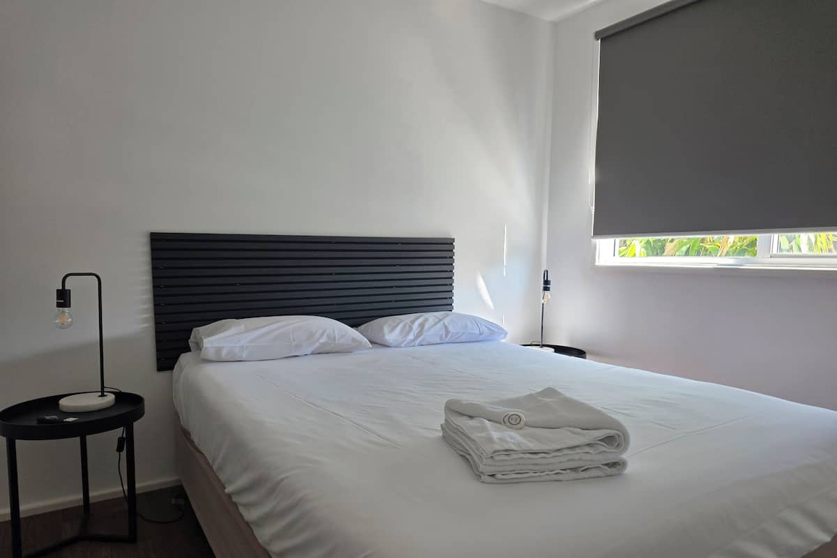 Una cama bien hecha con sábanas blancas, un cabecero negro y una ventana con vistas a la vegetación.