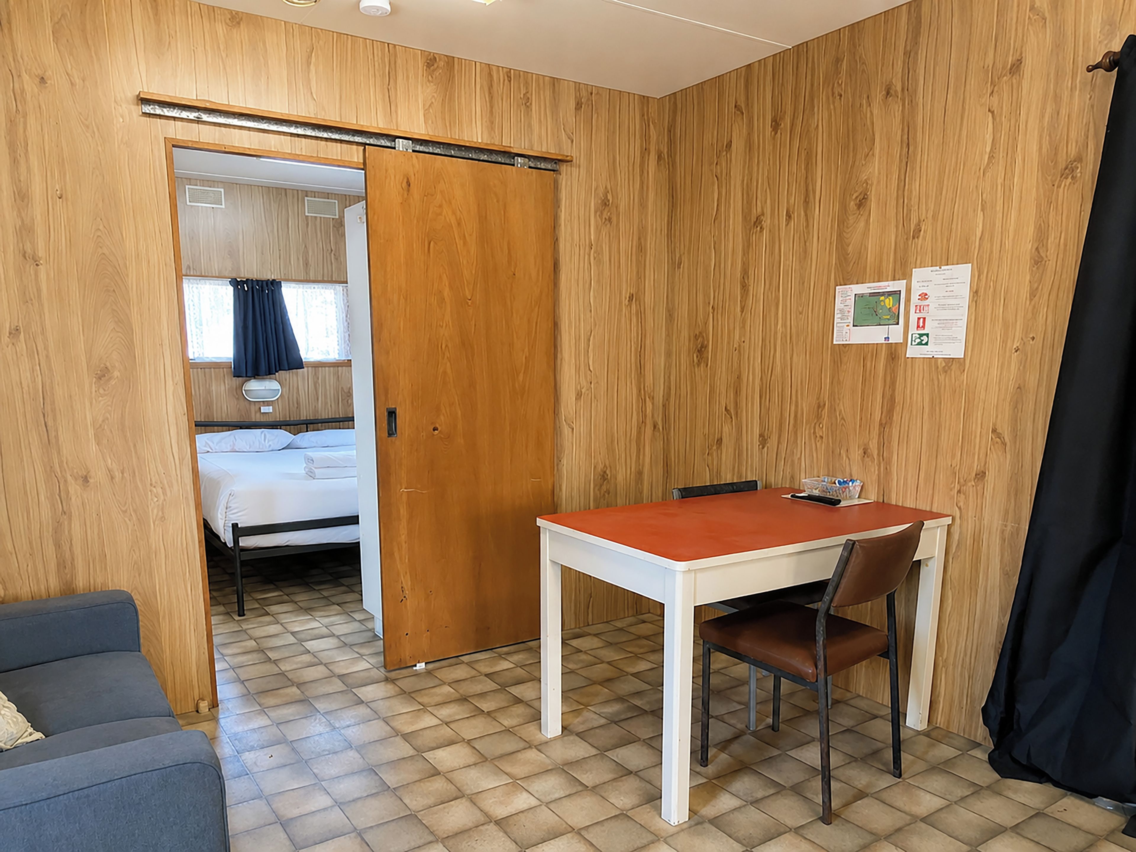 A room with a bed, a table with a chair, a sofa, and a wooden wall.