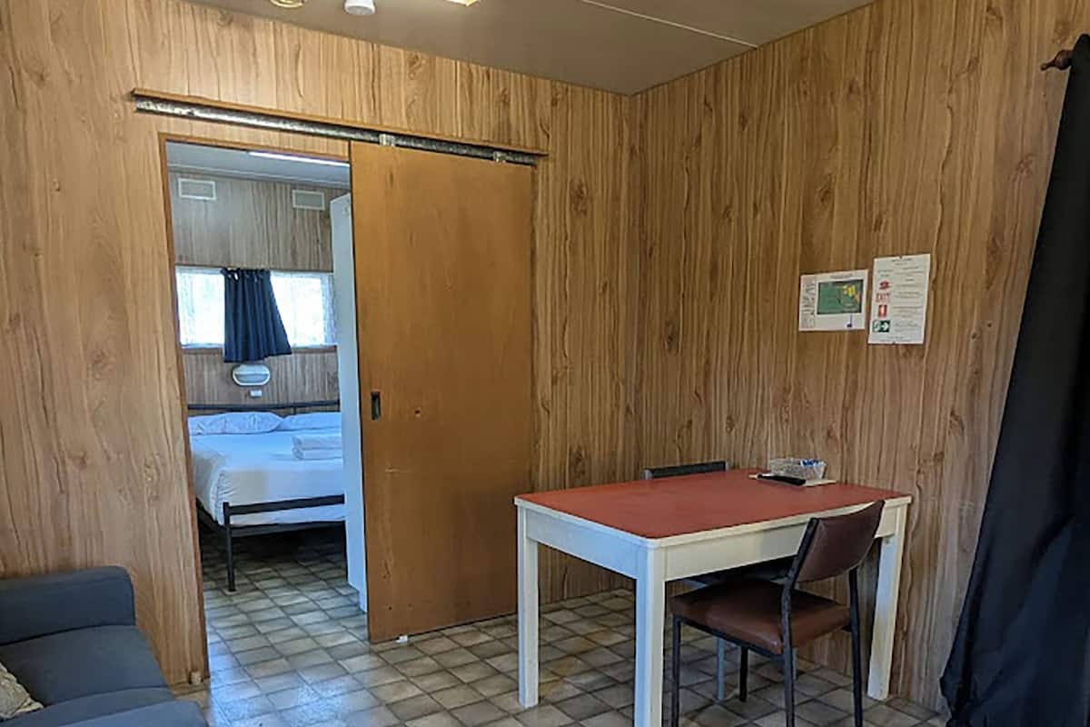 Una habitación con cama, una mesa con silla, una puerta de madera y piso de baldosas.