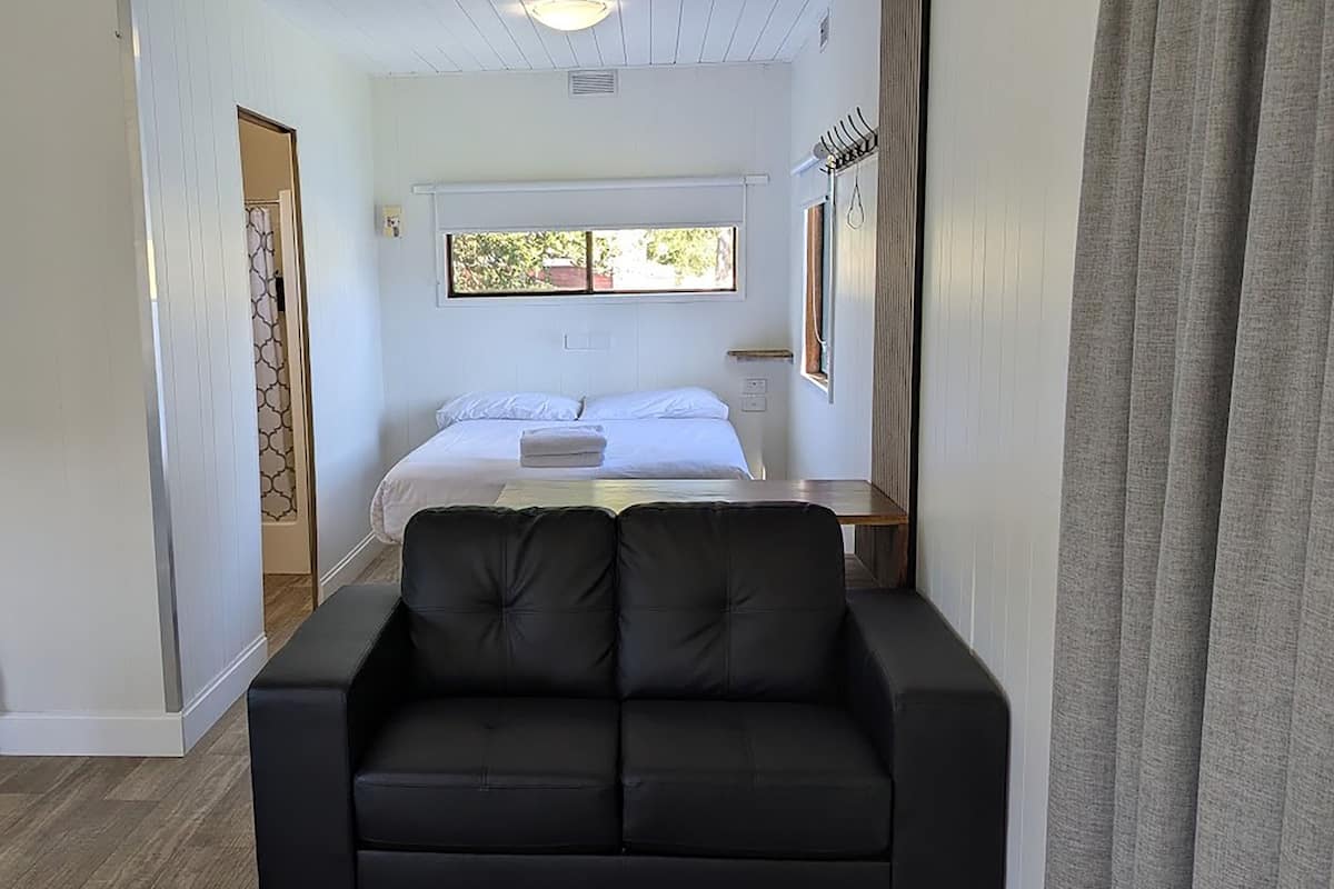 Una habitación con un sofá negro, una cama con ropa blanca, una ventana con vistas y un perchero.