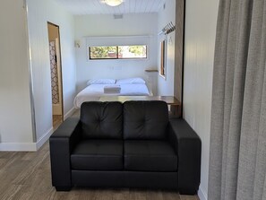 Una habitación con un sofá negro, una cama con ropa blanca, una ventana con vistas y un perchero.