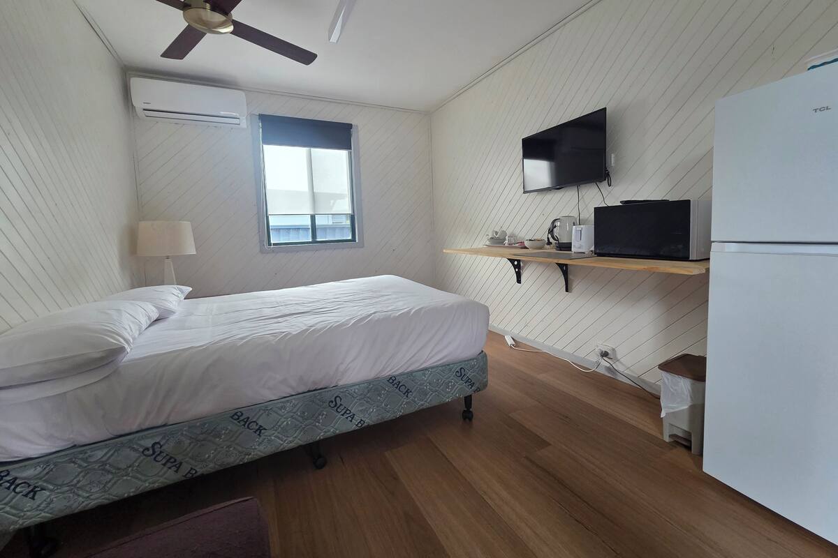 Un dormitorio con cama, ventilador de techo, aire acondicionado, ventana, televisor, microondas y refrigerador.