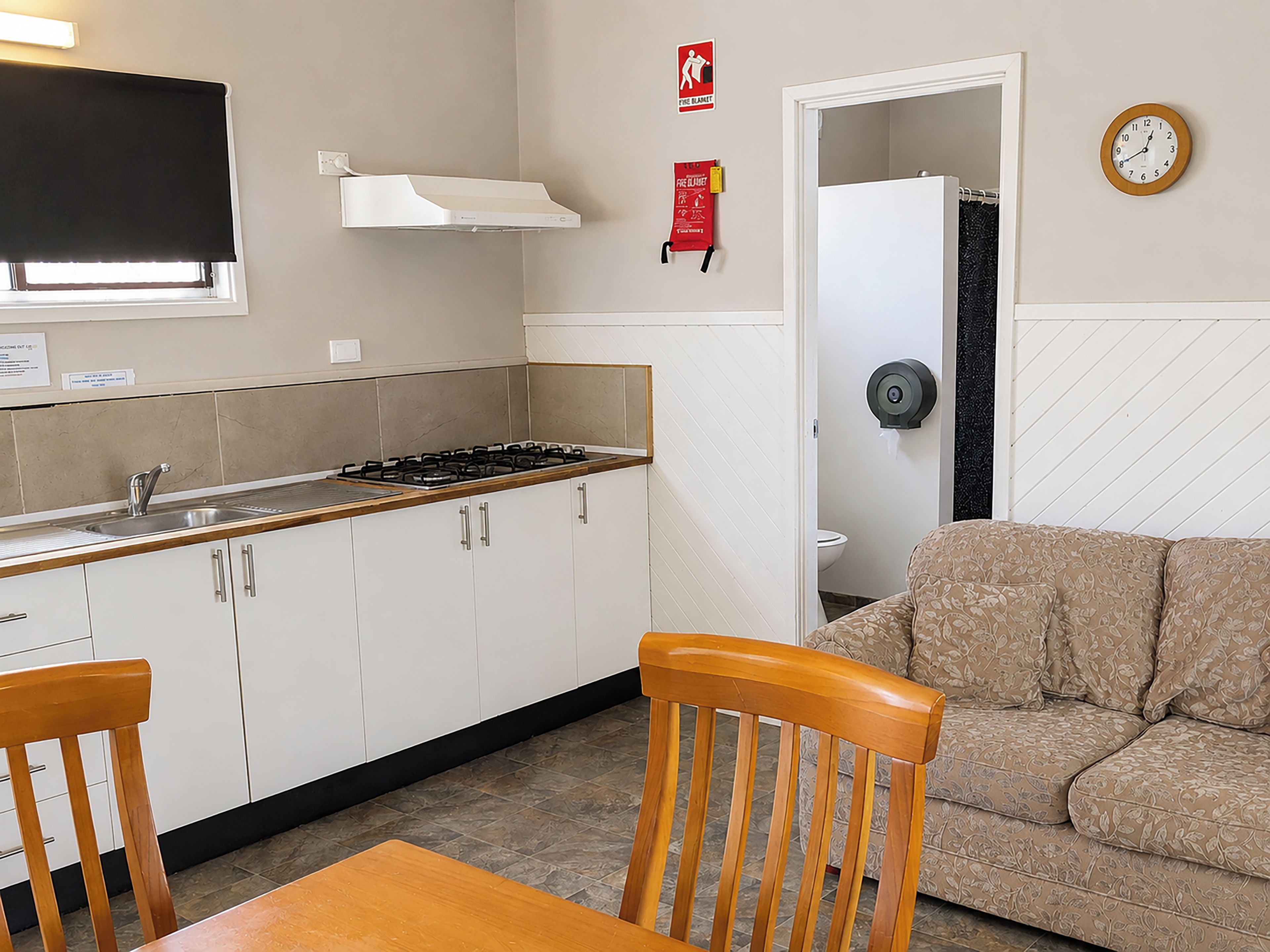 A kitchenette with a gas stove, cabinets, and a window with a blind. There is a sofa and a wooden table in the living area.