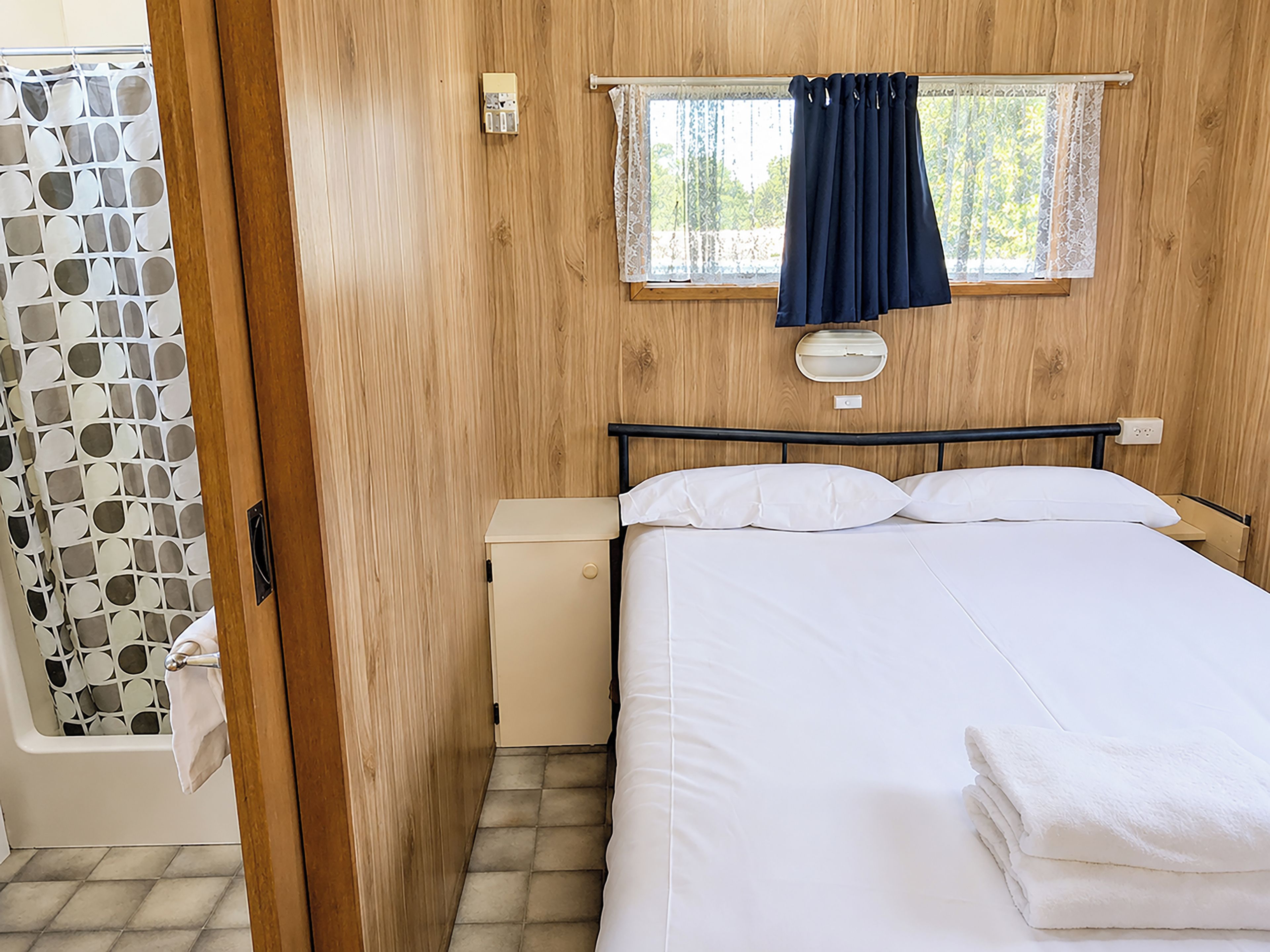 A bedroom with a bed, a nightstand, a window with curtains, a wooden wall, and a door.