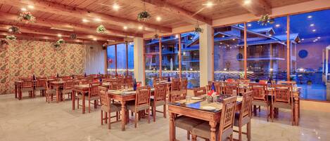 A spacious dining area with wooden tables and chairs, large windows offering views of the outside, and hanging plants.