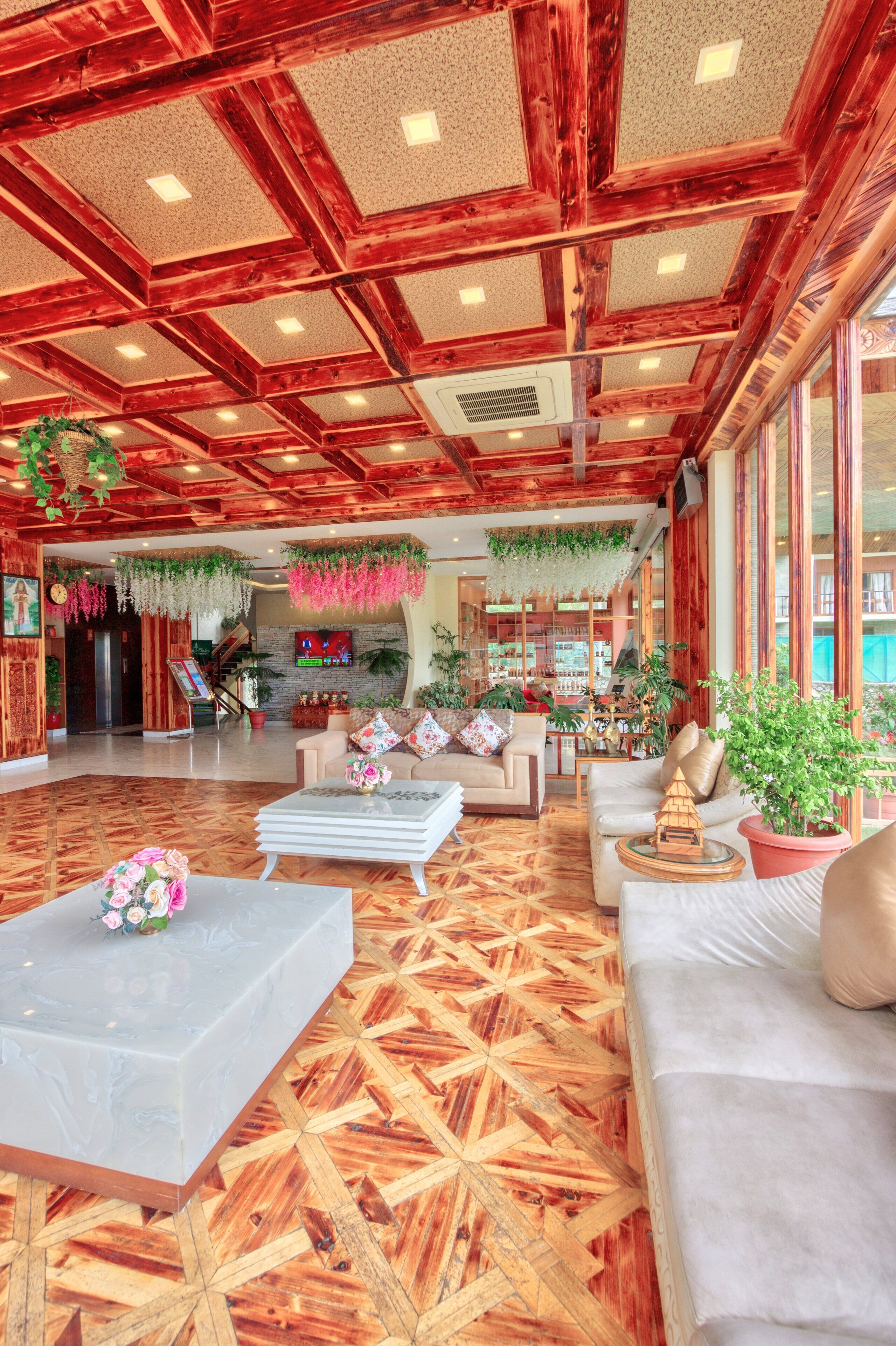 A spacious lobby with wooden flooring, a central seating area with sofas and a coffee table, and a bar area with plants and a television.