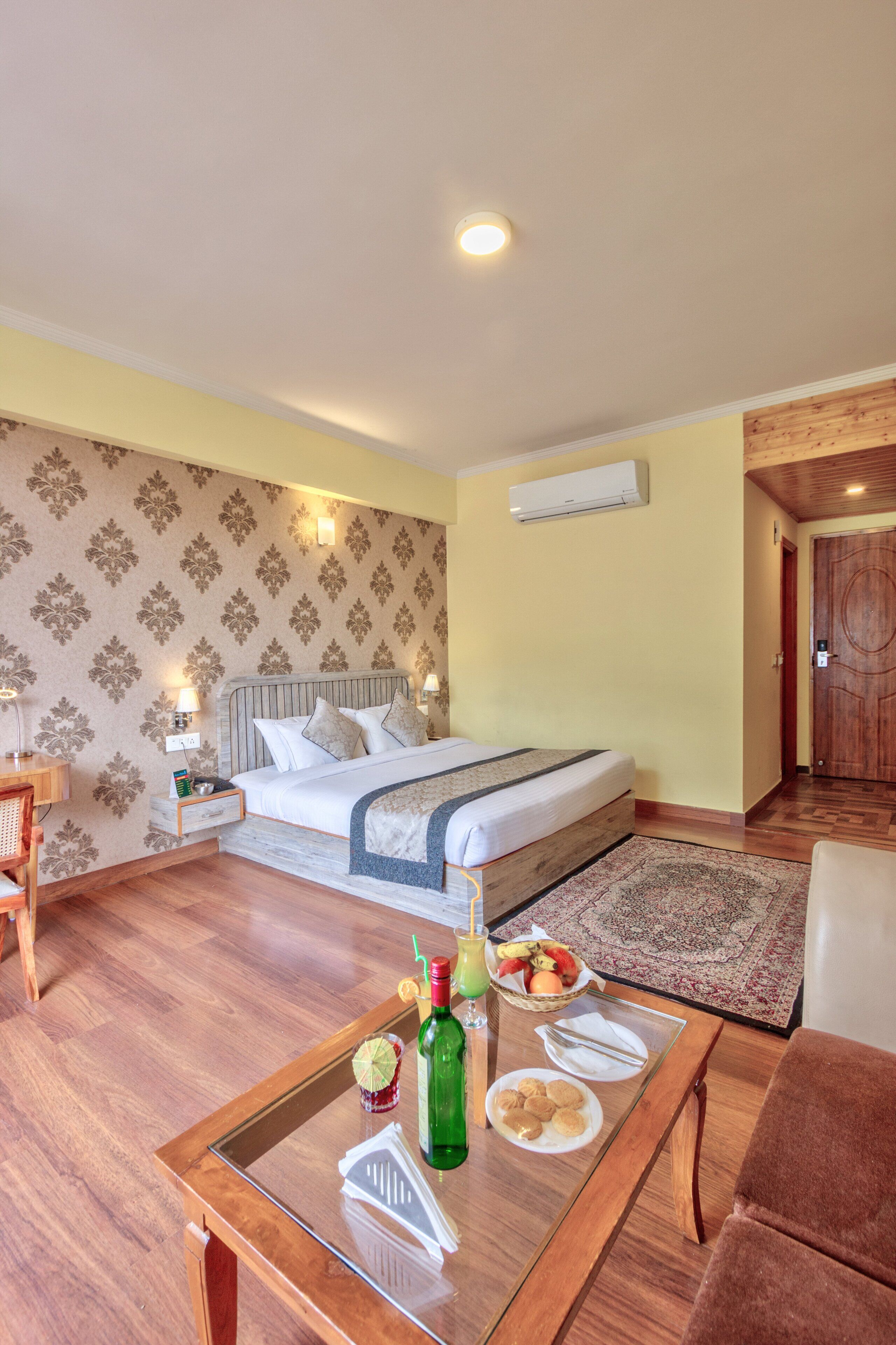 Quarto de hotel com cama, mesas de cabeceira, uma mesa de centro de vidro com itens de café da manhã, porta de madeira e papel de parede estampado.