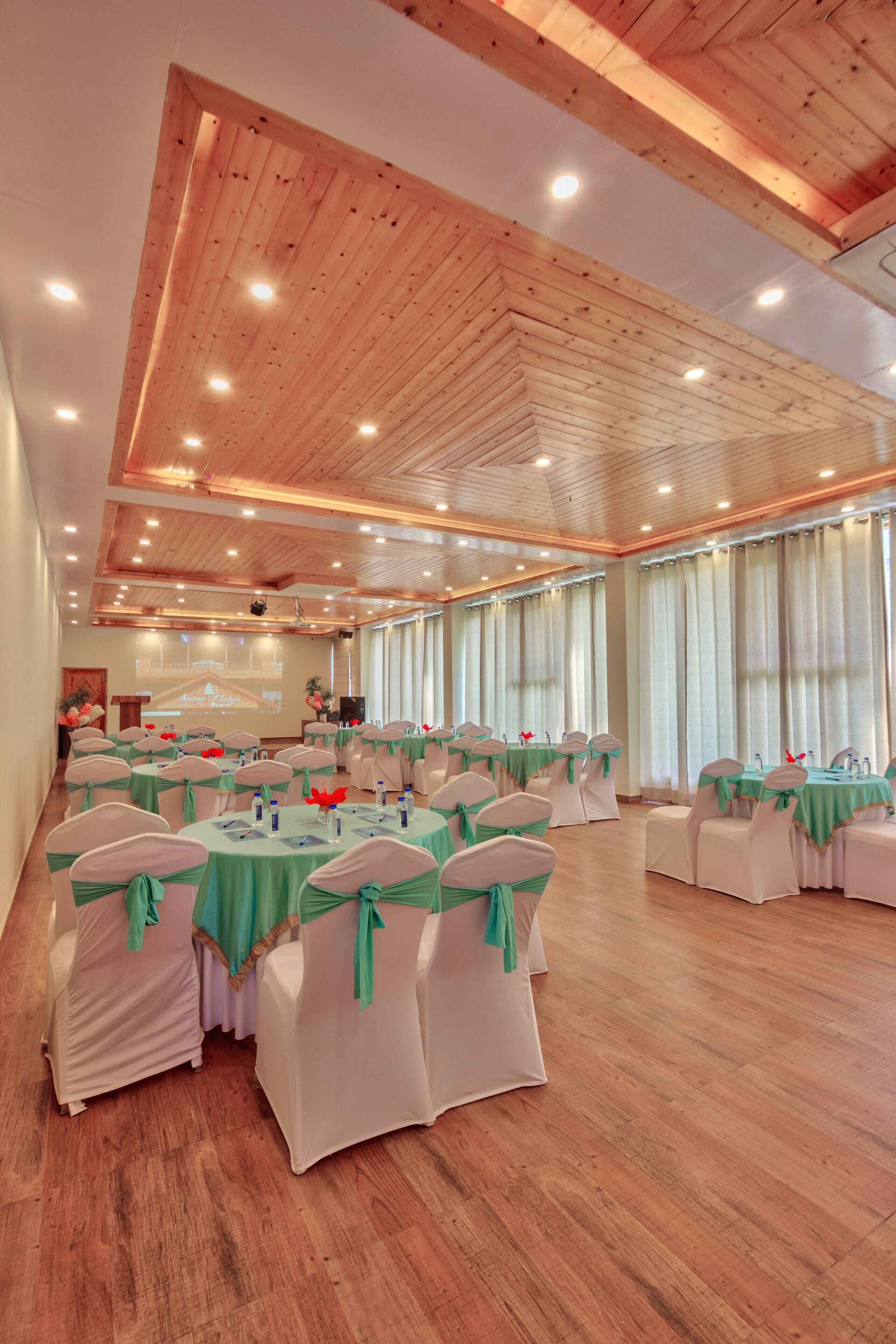 Une vaste salle dotée de plusieurs tables rondes, chacune recouverte d’une nappe verte et entourée de chaises blanches ornées de rubans verts. La pièce est pourvue d’un plancher en bois et d’un plafond haut avec des poutres en bois et un éclairage encastré.