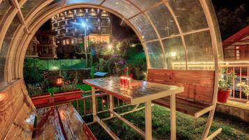 Un espace extérieur aménagé, abrité et doté de tables et de bancs, entouré de plantes en pot et offrant une vue sur un immeuble doté de nombreux balcons.