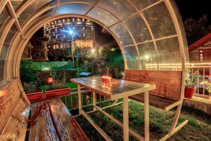 Un espace extérieur aménagé, abrité et doté de tables et de bancs, entouré de plantes en pot et offrant une vue sur un immeuble doté de nombreux balcons.