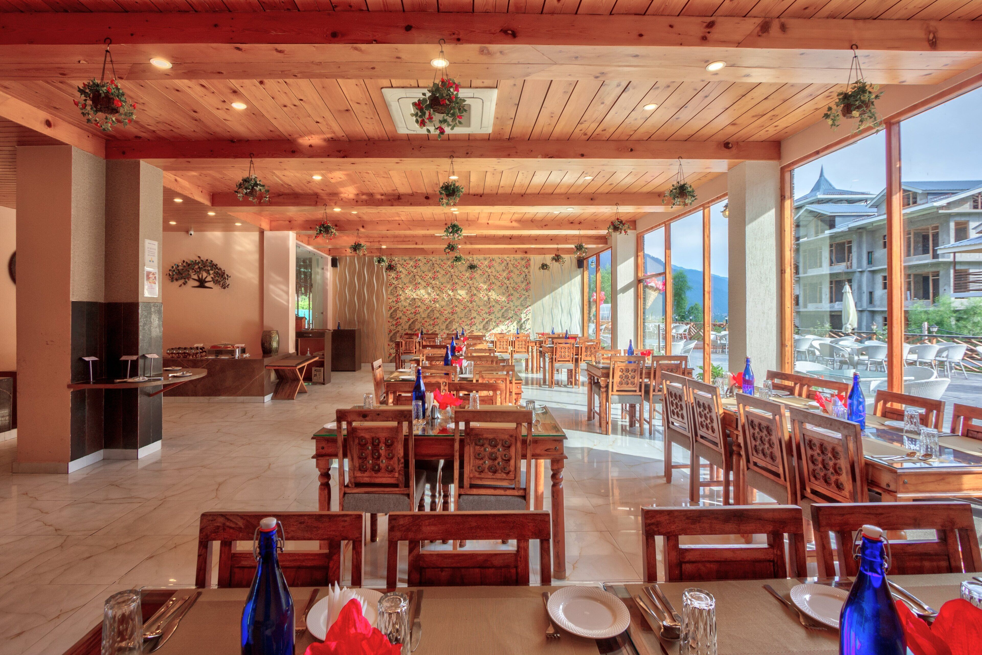 A spacious restaurant with wooden tables and chairs, large windows offering a view of the outside, and hanging plants.