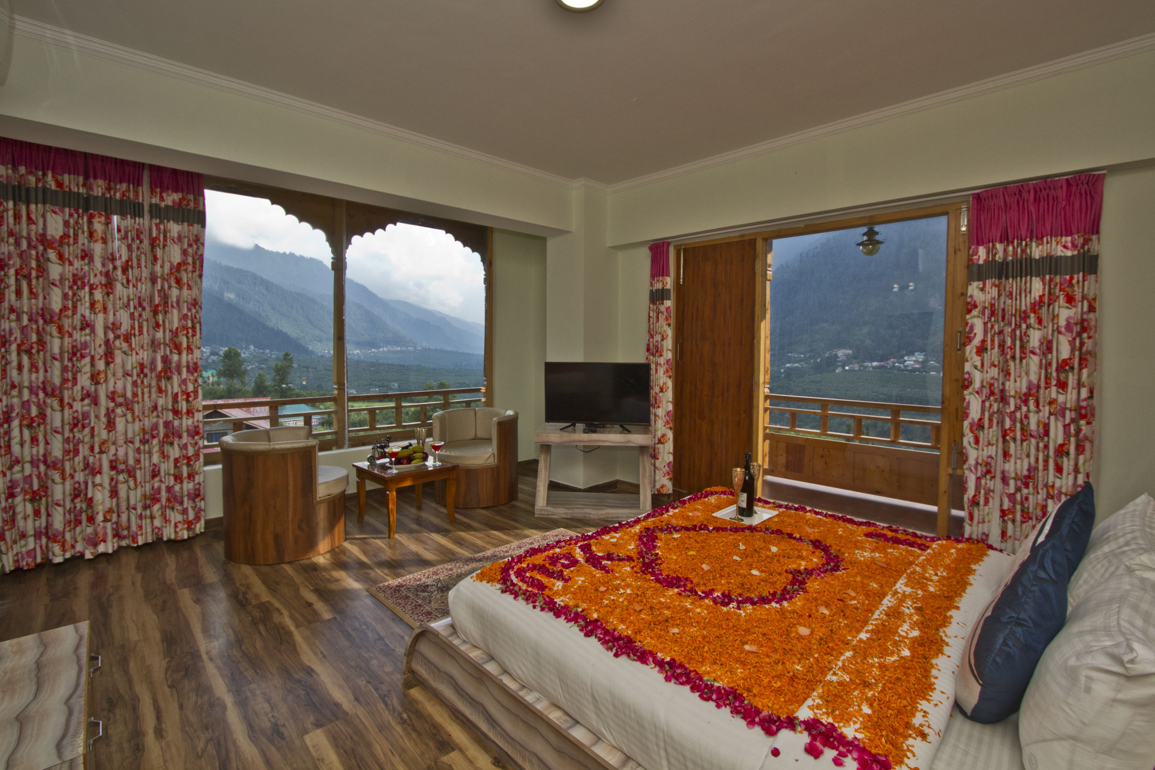 Habitación de hotel con una cama decorada con pétalos de flores, un escritorio con televisor y un balcón con vista a la montaña.