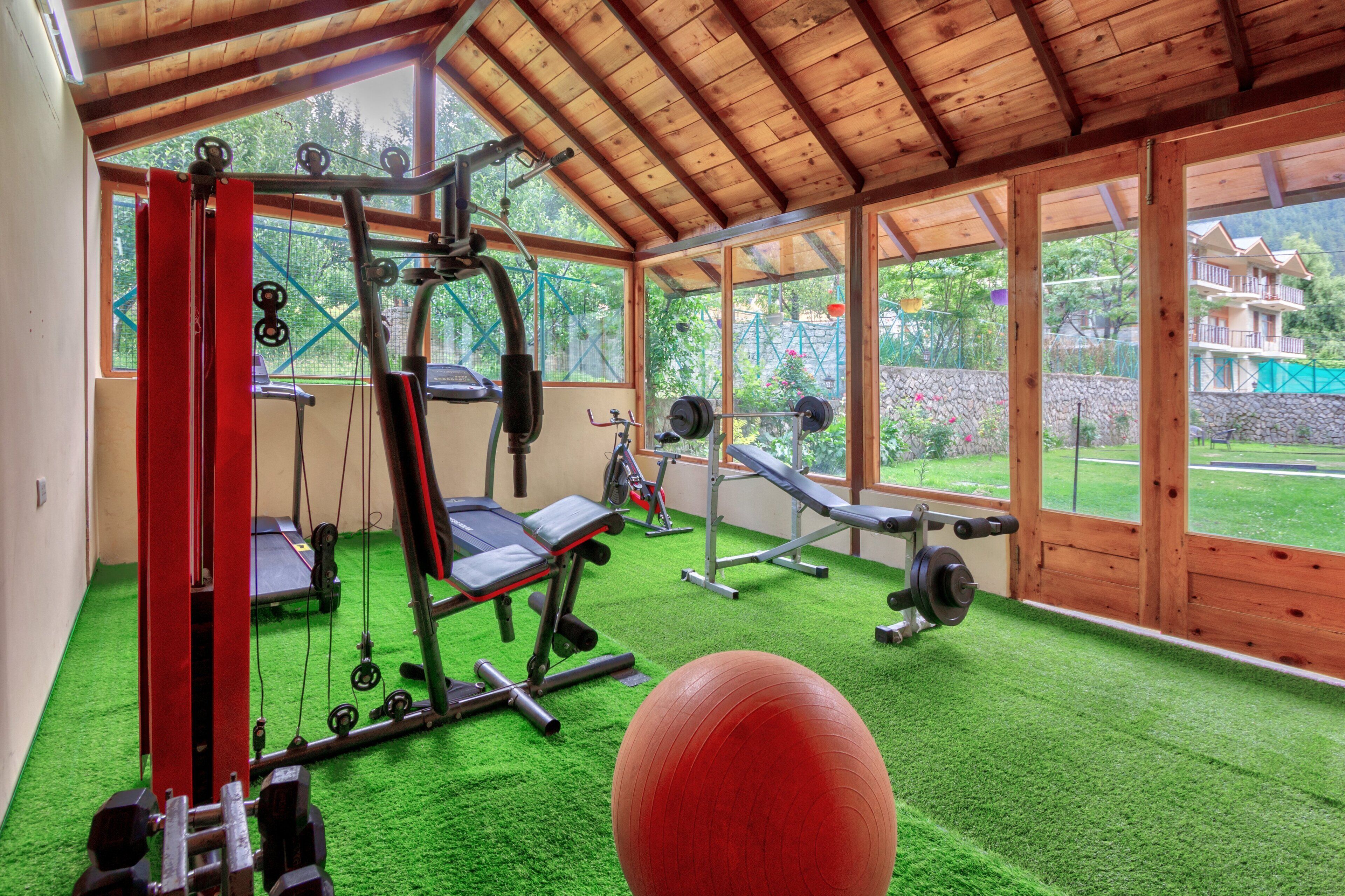 Academia com equipamentos de exercício, uma bola vermelha de ginástica e vista para um campo verde e edifícios através de grandes janelas.