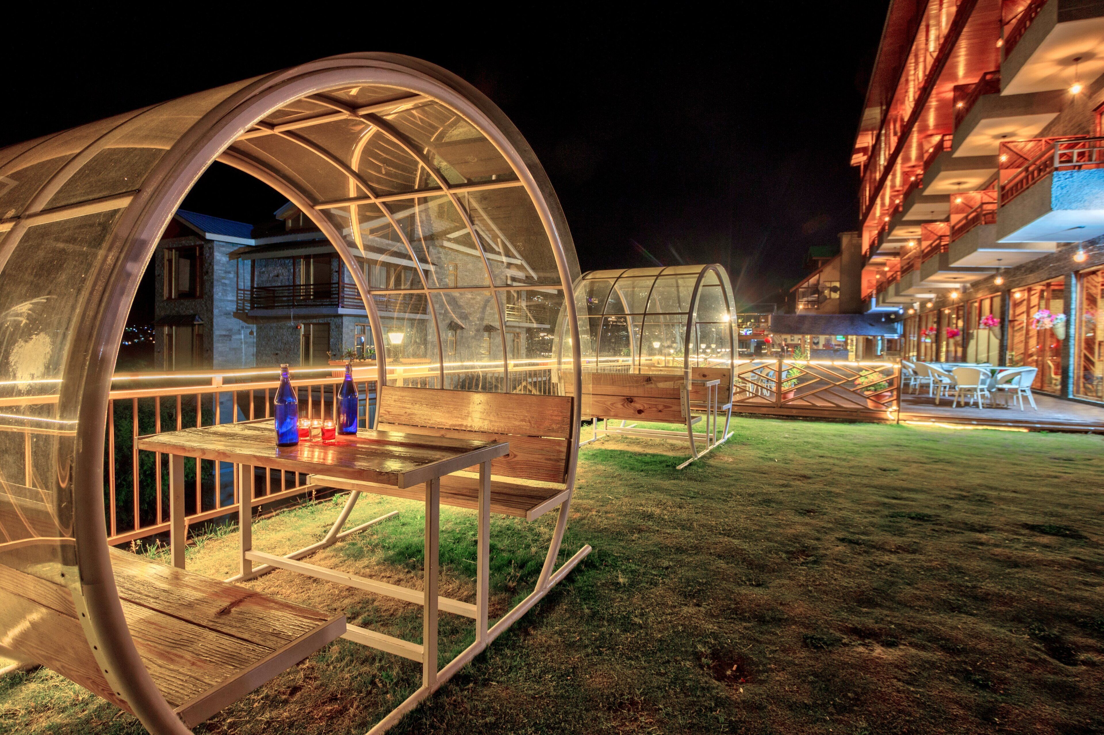 Une structure en forme de dôme transparent, abritant une table et des bancs, entourée d’une pelouse et d’un bâtiment doté de balcons.