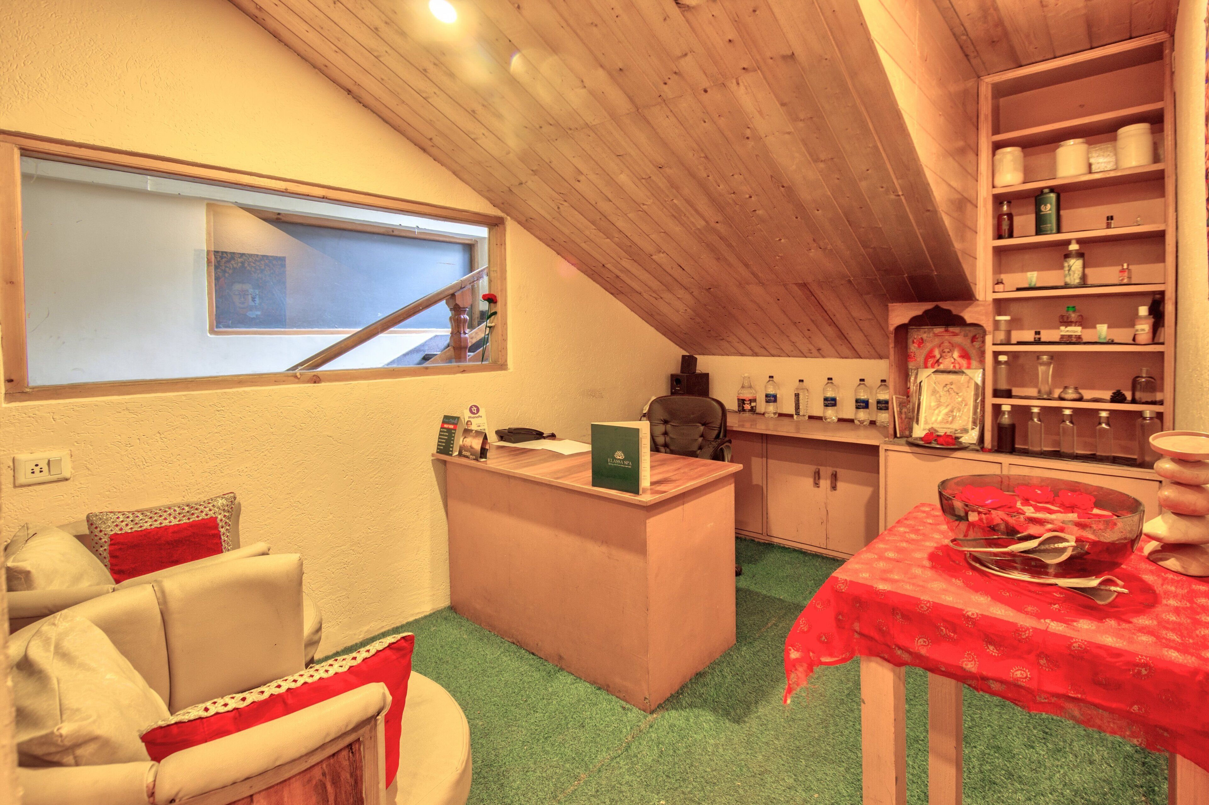 A cozy attic room with a wooden desk, a chair, a small couch, a dining table with a red tablecloth, and a window with a view.