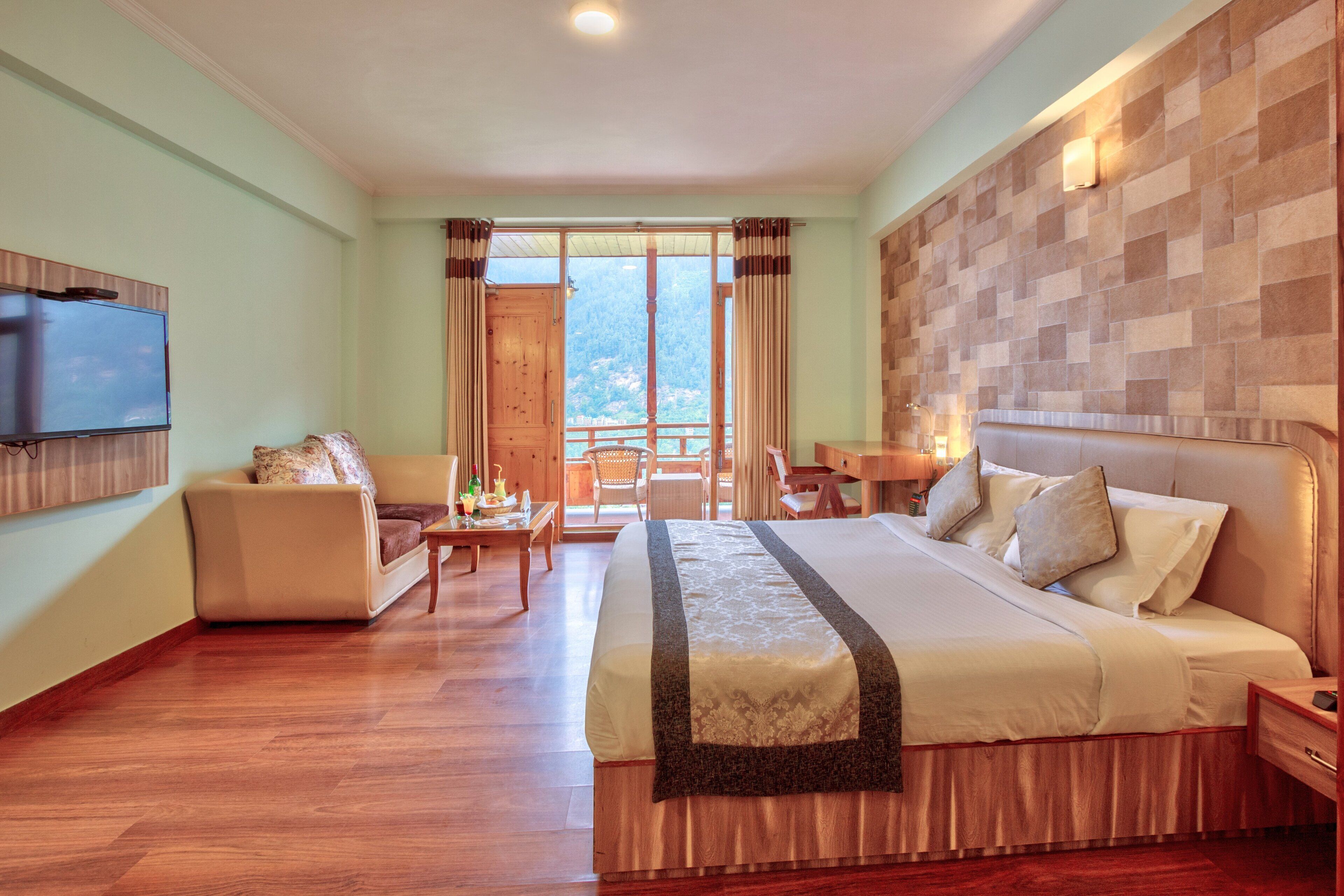 A hotel room with a large bed, a TV, a balcony with a view, and a wooden floor.