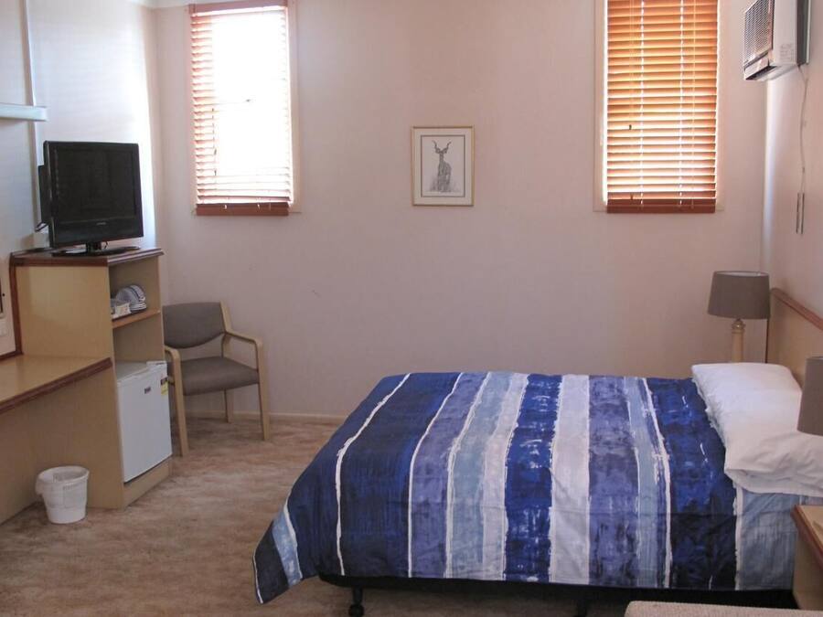 Une chambre à coucher comprenant un lit, un bureau, une chaise, un téléviseur et une fenêtre avec des stores.