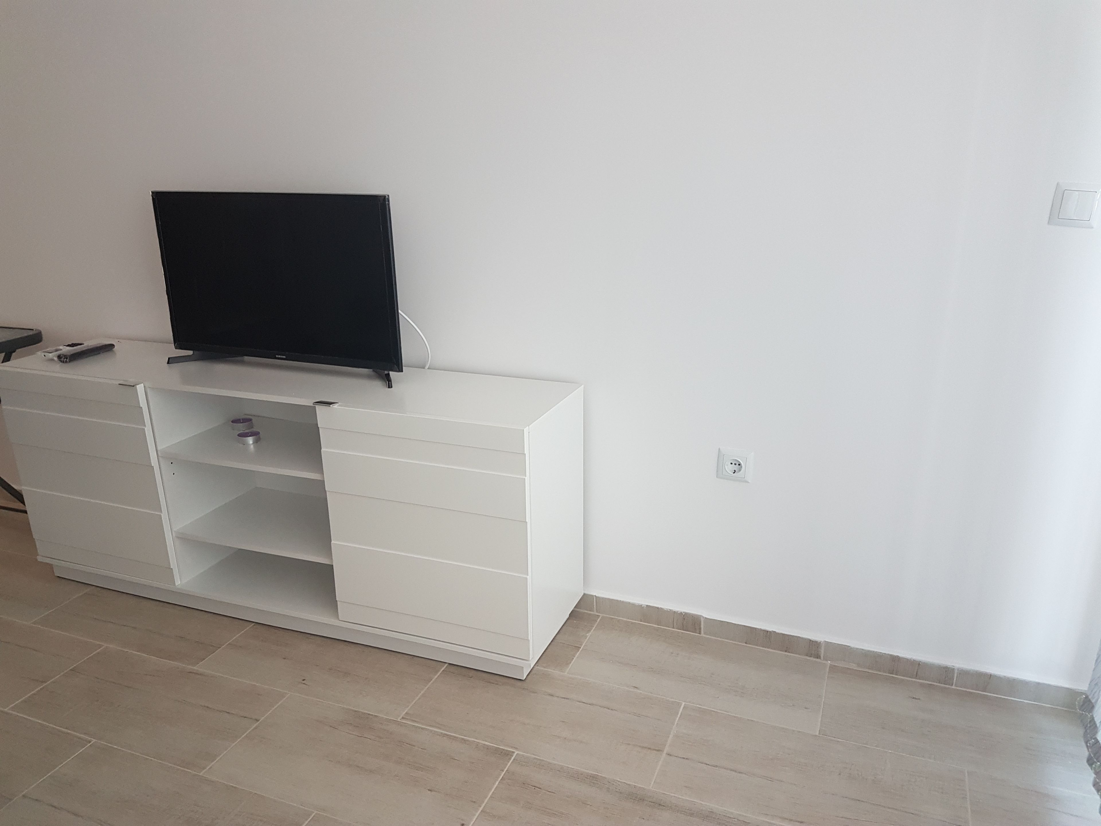 A white entertainment unit with a flat-screen TV, a remote, and a small object on the top shelf.