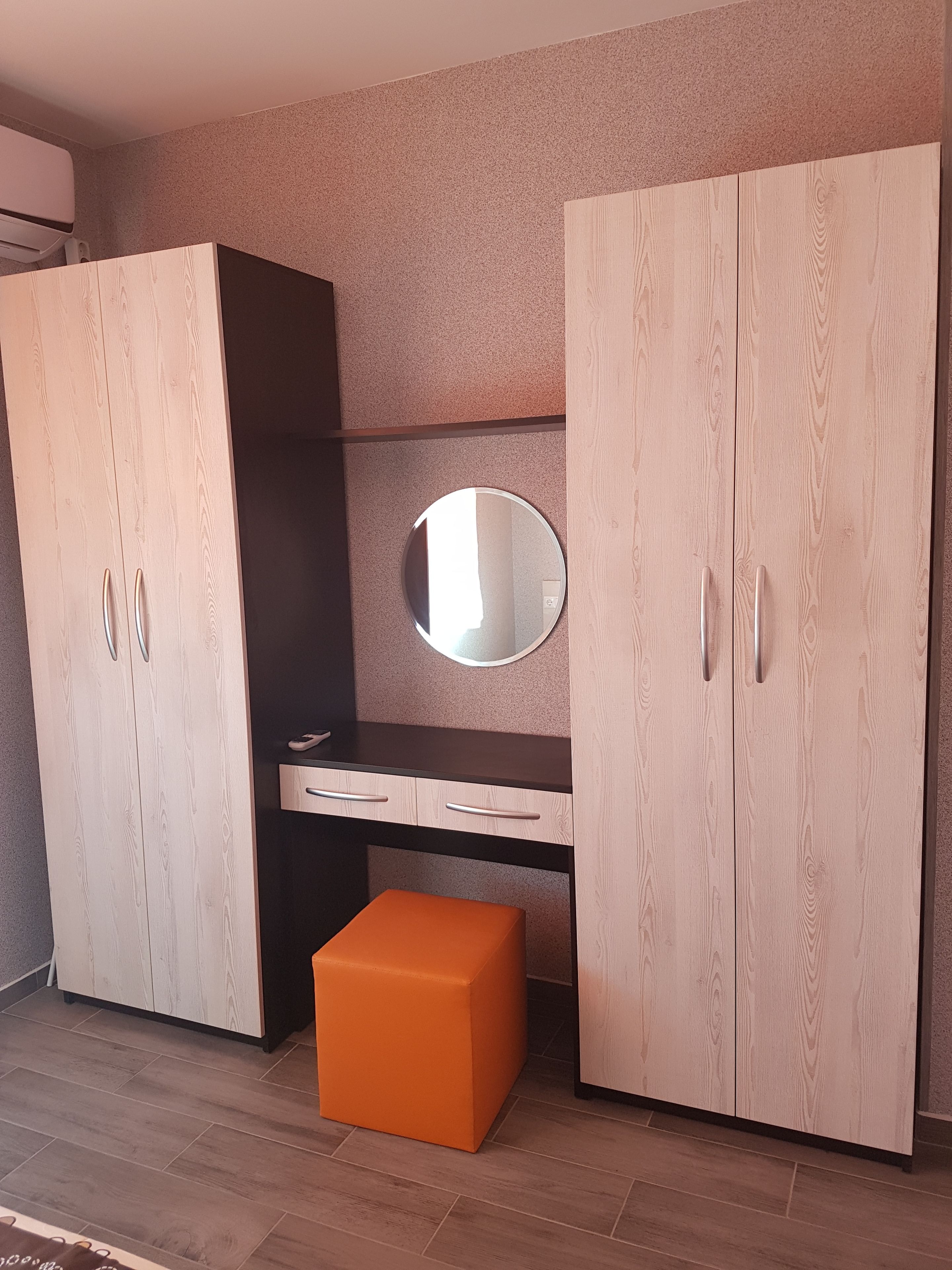 A room with a wardrobe, a mirror, a desk, and an orange ottoman.
