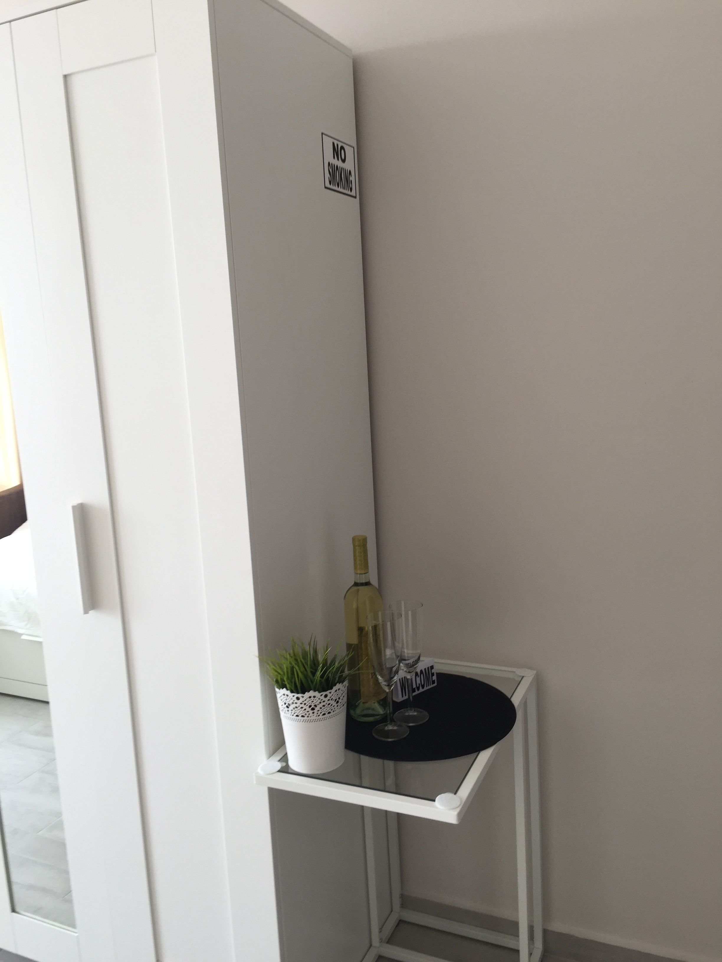 A corner of a room with a white door, a 'NO SMOKING' sign, a small white table with a potted plant, a bottle of wine, and two glasses.