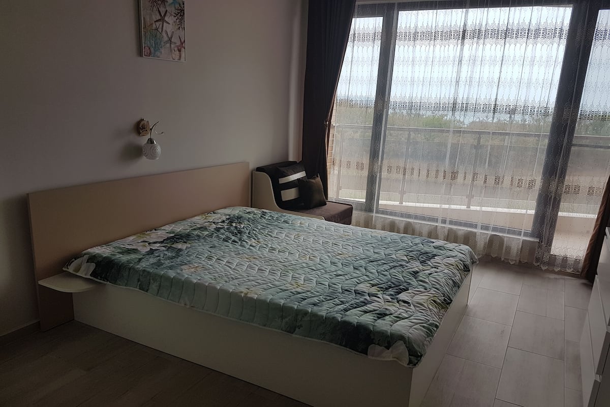 A bedroom with a bed, a chair, a small table, a window with curtains, and a wall decoration.