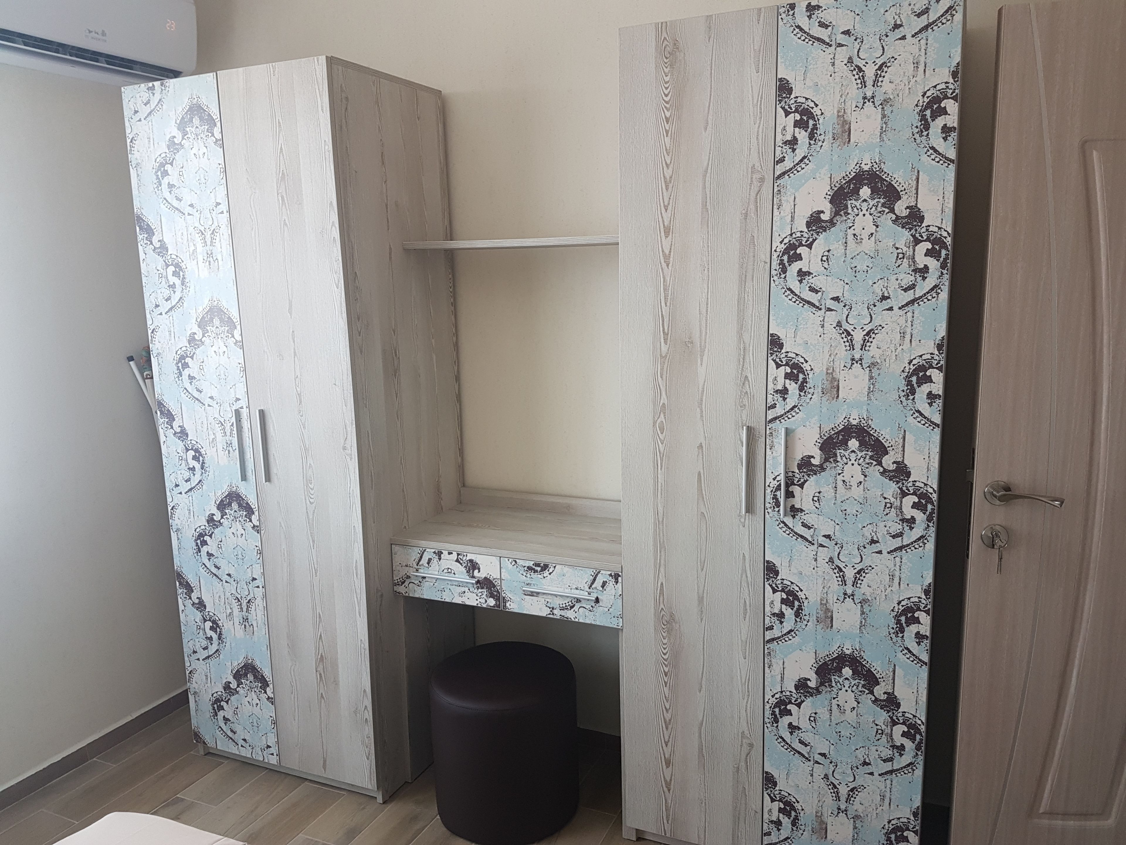 A room with a wardrobe featuring a light wood finish and patterned doors, a small stool, and a shelf unit.